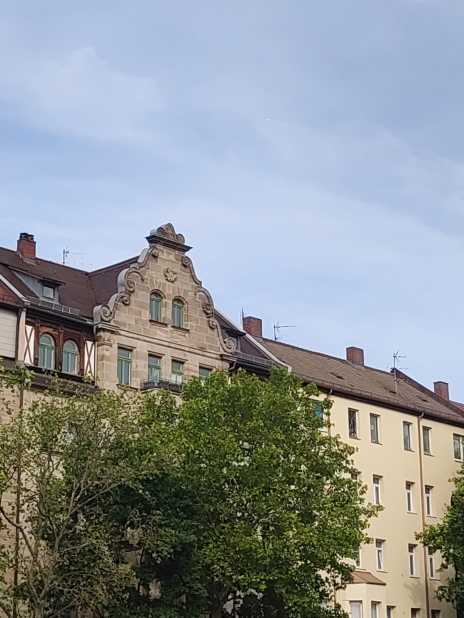 The image depicts an urban scene in Fürth, Germany, focusing on the facades of buildings and the sky. In the foreground, lush green trees obscure parts of the lower sections of the buildings. The architecture includes a historical building with a decorative stone gable, featuring scrollwork and a central emblem, alongside a more modern, pale yellow apartment building. Both buildings have tiled roofs with chimneys and visible antennas. Windows of various shapes and sizes are present on both structures, some with green frames, others with white. The sky above is a pale blue with scattered clouds, and a faint contrail from an aircraft is visible high up. The overall impression is a daytime view of residential and historic architecture in a German city.