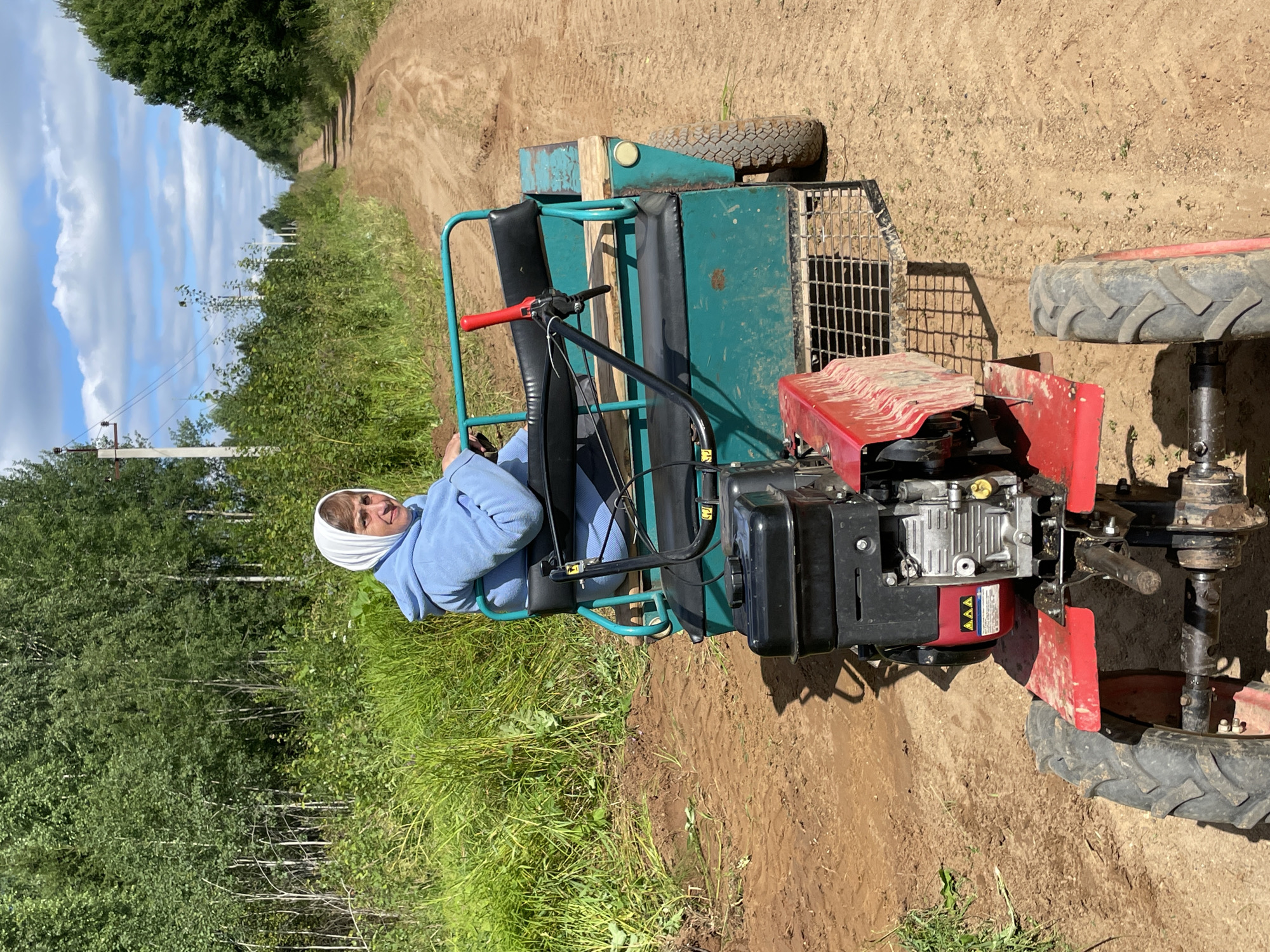 A woman wearing a light blue hooded sweatshirt and a white headscarf sits on a green trailer attached to a red and black tiller. The tiller has a prominent engine and two large wheels. The trailer has two seats, the one the woman is occupying is black and padded, and there is a red lever on the side. The trailer also has a green metal frame. The tiller and trailer are on a dirt road. Tall green grass and trees line the sides of the road. The sky is blue with white clouds. The location is identified as Dresvyanovo, Russia.