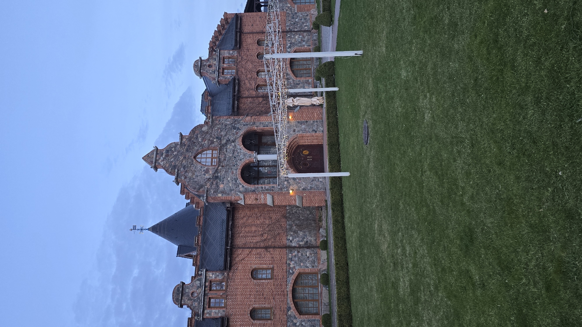 The image depicts a building with castle-like architecture located in Kaliningrad, Russia. The building is constructed from red brick and gray stone. It has multiple windows and arched doorways. A grassy lawn is adjacent to the building, and part of the sky is visible in the upper left corner.