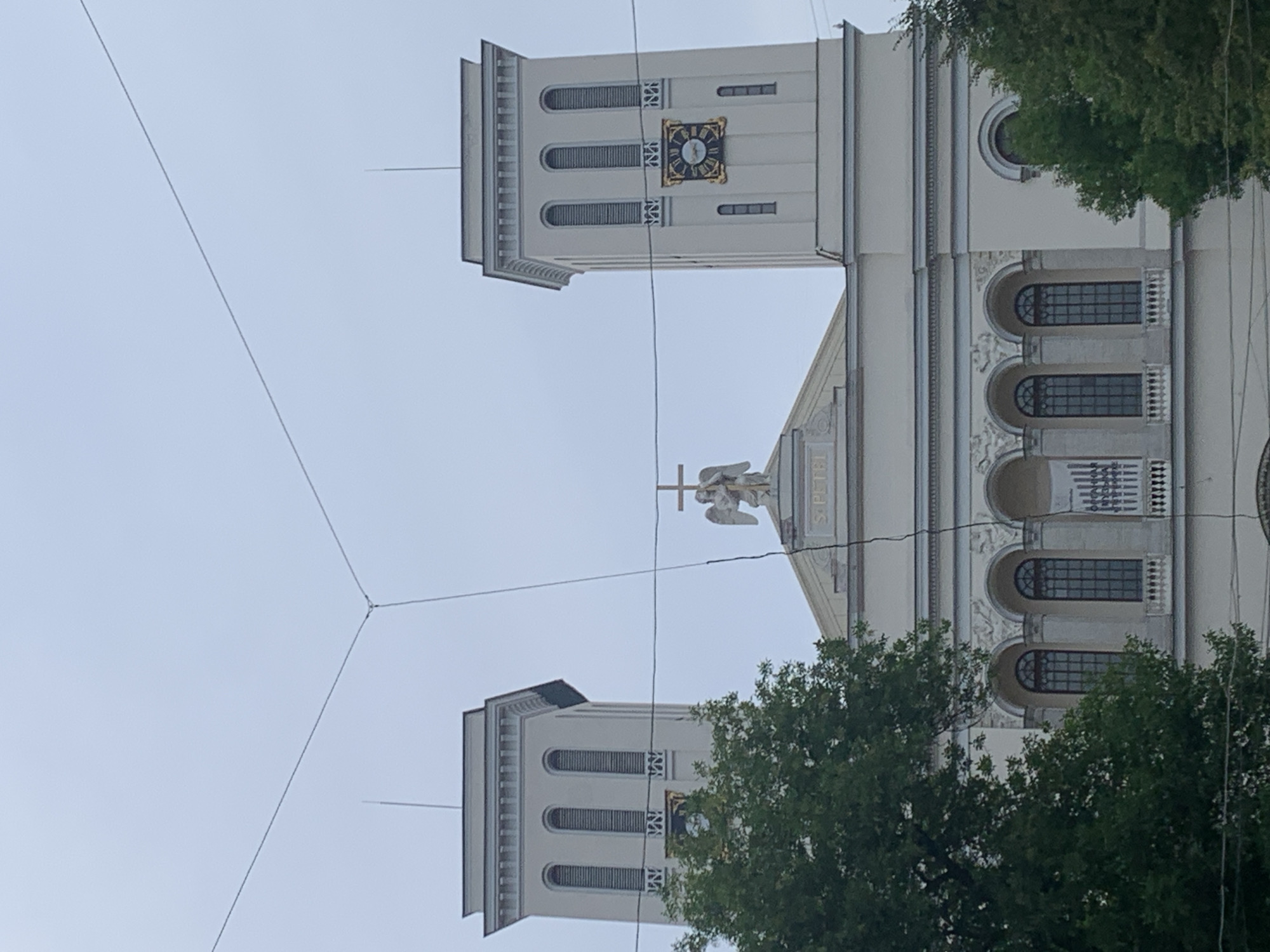 This is an upright, eye-level, exterior shot of the Saint Petersburg Catholic Church of the Assumption of the Virgin Mary. The church is a neoclassical building with two prominent towers. The left tower features arched windows and a clock face. The right tower is partially obscured by trees, but it also has windows. Atop the central section of the building, between the towers, is a statue of an angel holding a cross. The sky above is overcast and a pale blue. There are power lines crisscrossing the foreground. The image appears to have been taken during the daytime. The text "S. PETR" is visible on a banner above the angel statue.