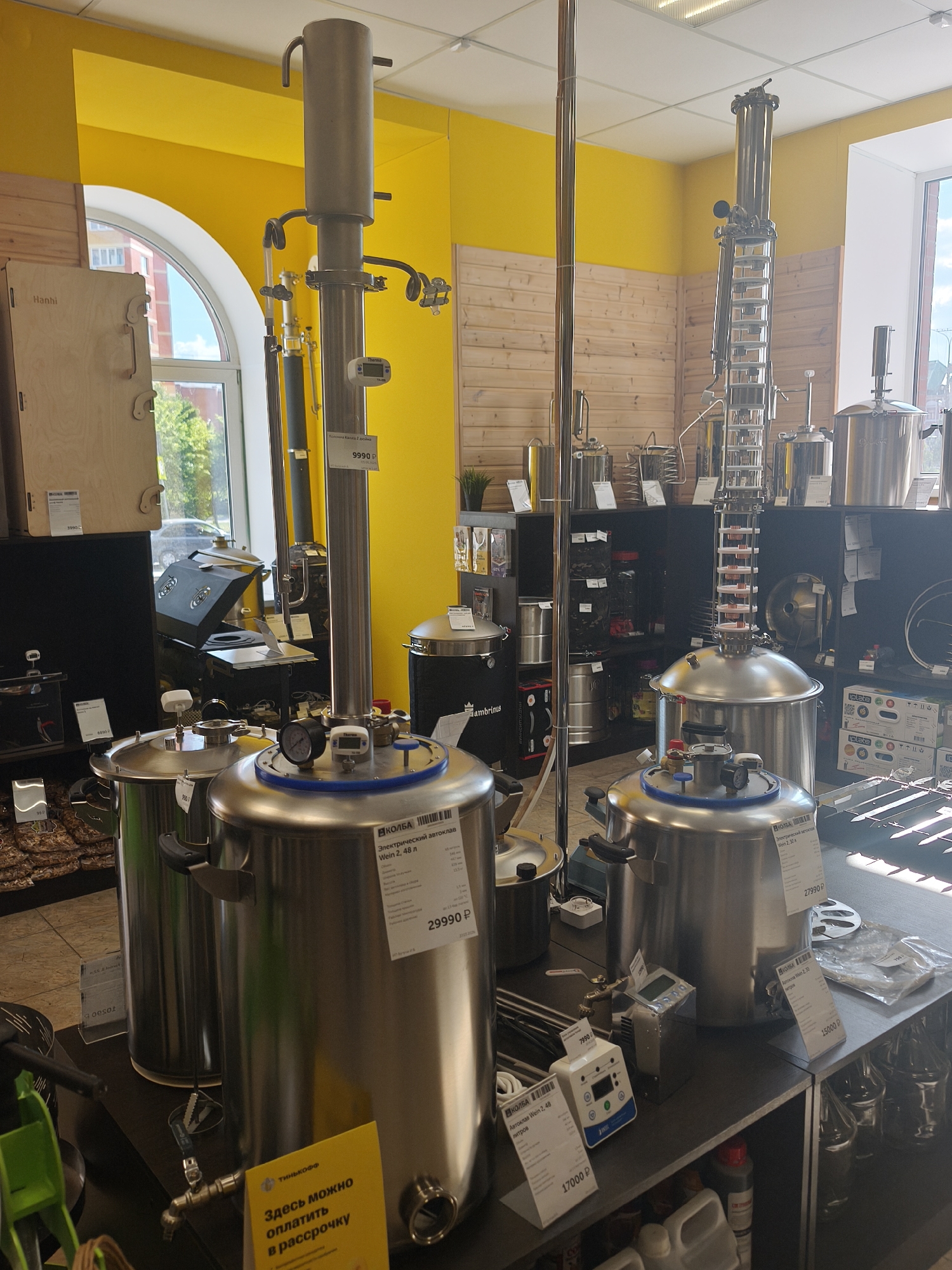 The image depicts the interior of a store selling distillation equipment and related items. Several large stainless steel stills of various sizes are displayed on shelves and tables. The store features a bright yellow wall and light-colored wooden paneling. Various price tags and informational labels are visible on the products. The overall impression is that of a retail environment specializing in brewing or distilling apparatus. The location is confirmed as Yoshkar-Ola, Russia, based on the presence of Russian text on some of the labels and the likely context of the establishment within that city.