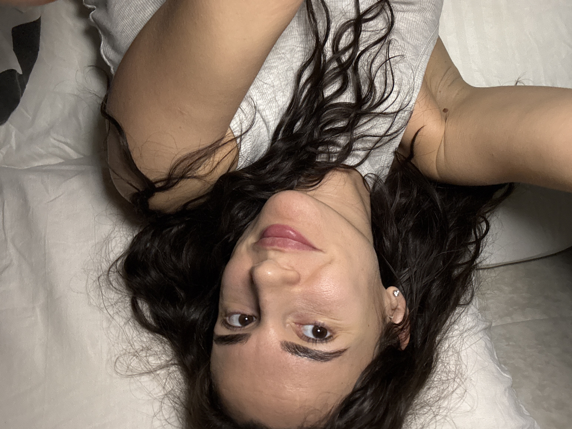 A woman with long, dark, wavy hair lies on a white surface, possibly a bed or examination table. She is wearing a light gray top. Her head is positioned at an angle, with her face turned towards the viewer. She has fair skin, dark eyebrows, brown eyes, and pink lips. A small earring is visible in her left ear. The image's perspective suggests it may be a selfie taken from above. There is no definitive indication of the location being Chaikovsky, Russia.