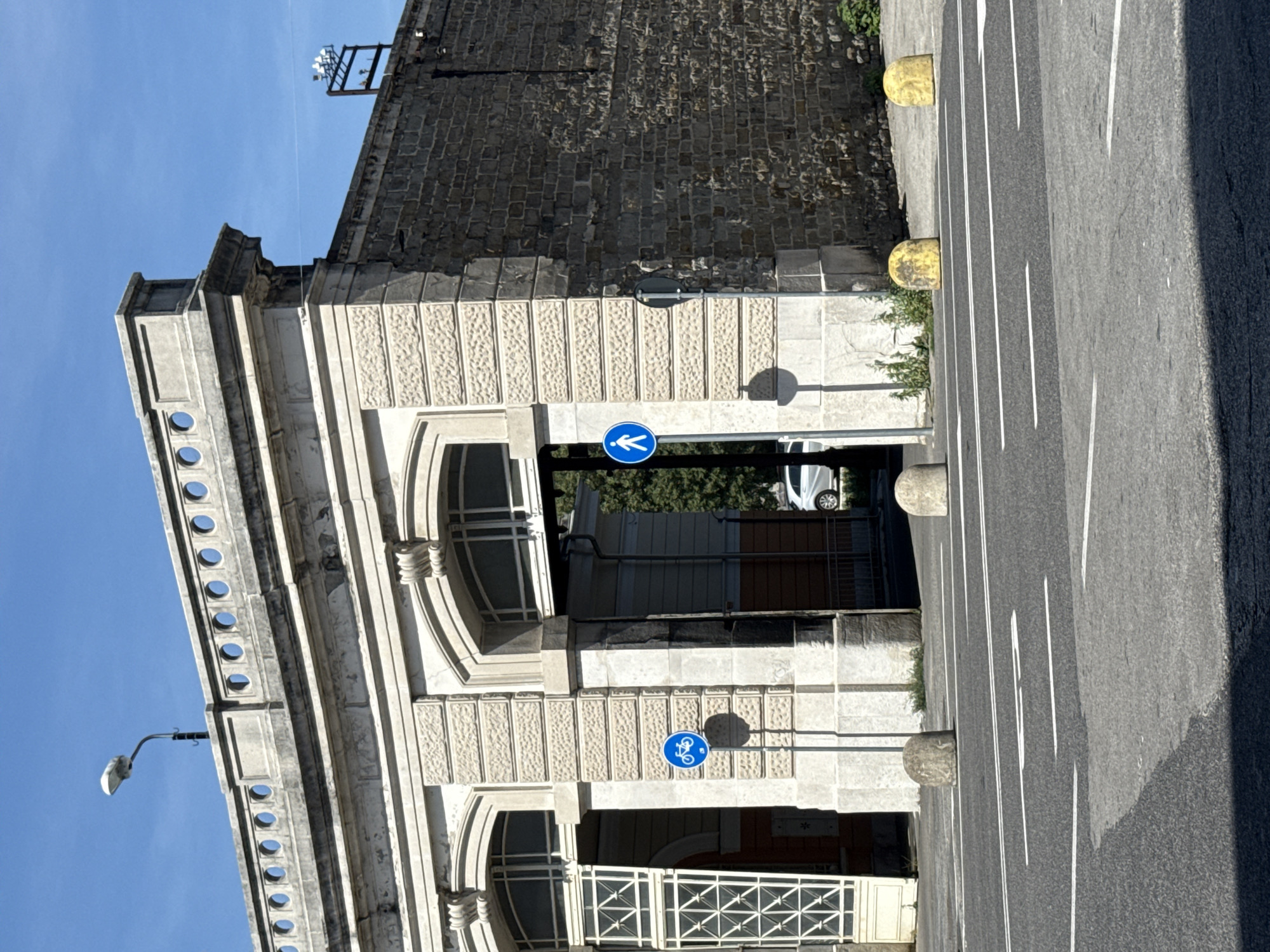 The image shows a view of a building in Trieste, Italy, with a street in the foreground. The building is constructed of stone and features large archways. A street lamp is visible on top of the building, and a sign indicating a bicycle path is posted on a pole to the left. Another sign indicating a pedestrian path is posted on a pole to the right. There are several round bollards on the sidewalk. The street is paved and marked with white lines. The sky is clear and blue.
