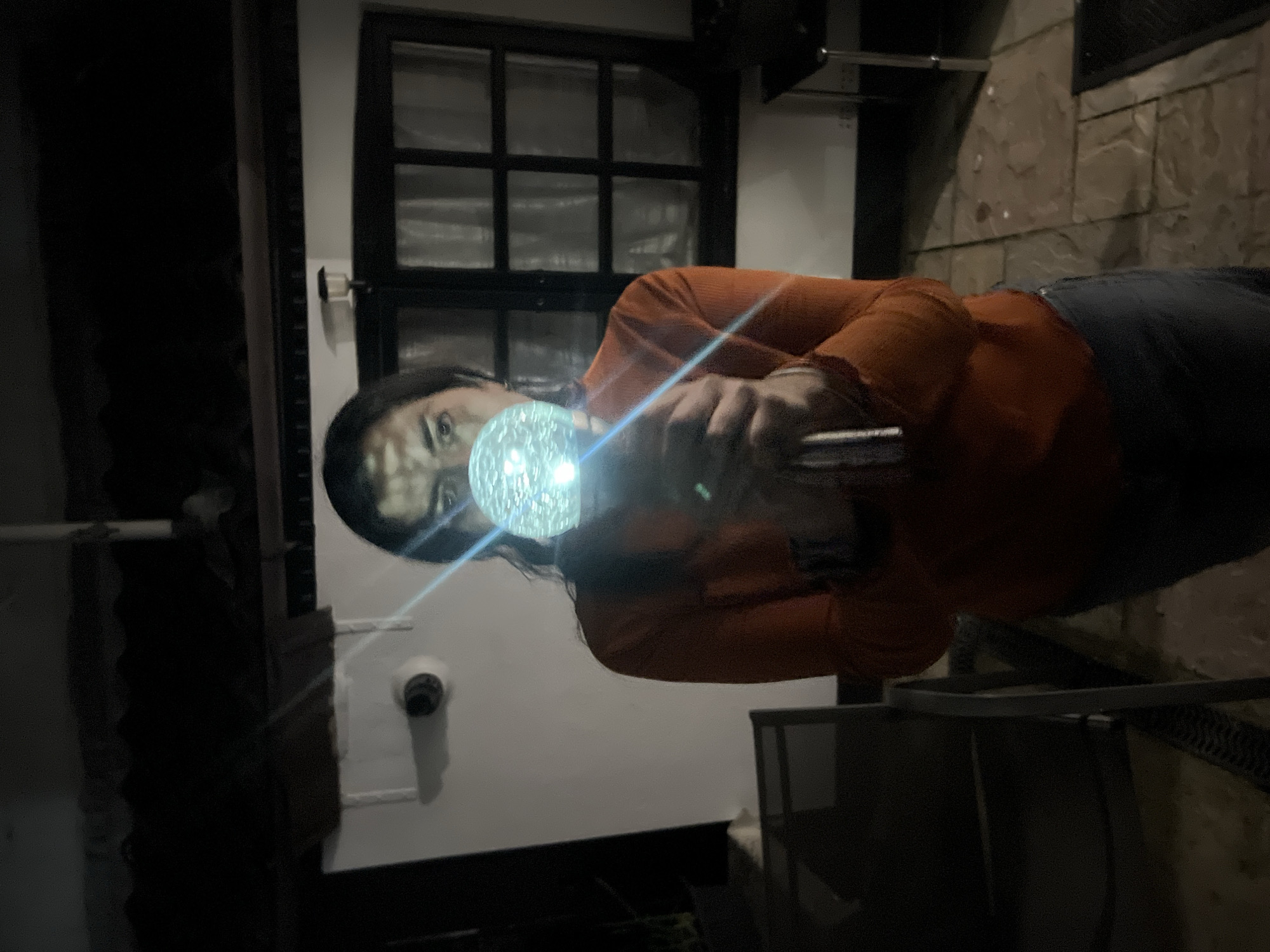 A person is standing outside in the dark. They are holding a brightly lit, spherical object directly in front of their face. The person is wearing an orange long-sleeved shirt and dark pants. Behind the person is a building with a window composed of multiple panes. To the right, there is what appears to be a patio area with chairs and a stone or brick surface. The exact location within Gorleston-on-Sea is not discernible from the image.