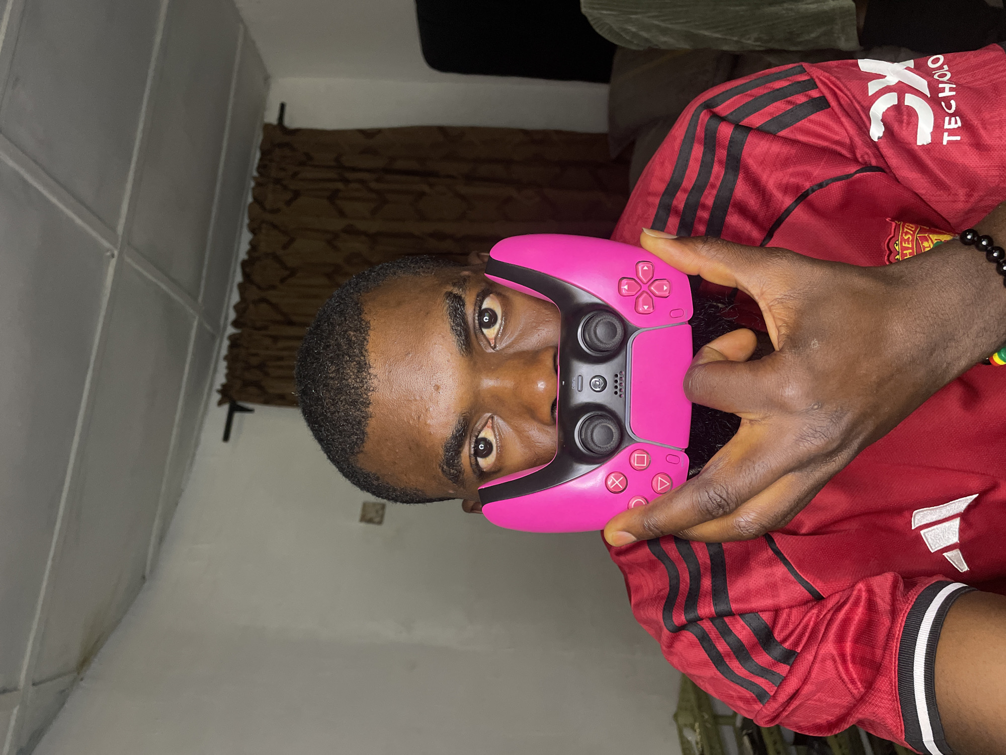 A medium shot shows a Black man in a red Manchester United jersey, holding a pink video game controller to his face. The controller covers his mouth and nose, obscuring his lower face. His eyes are visible, looking directly at the camera. The man has dark skin, short dark hair, and a trimmed beard. The background shows a plain white wall, brown curtains, and a white ceiling with visible panels. The scene is lit from above, casting subtle shadows. The image appears to have been taken indoors, possibly in a residential setting. There are no visible indications of the specific location within Jos, Nigeria.