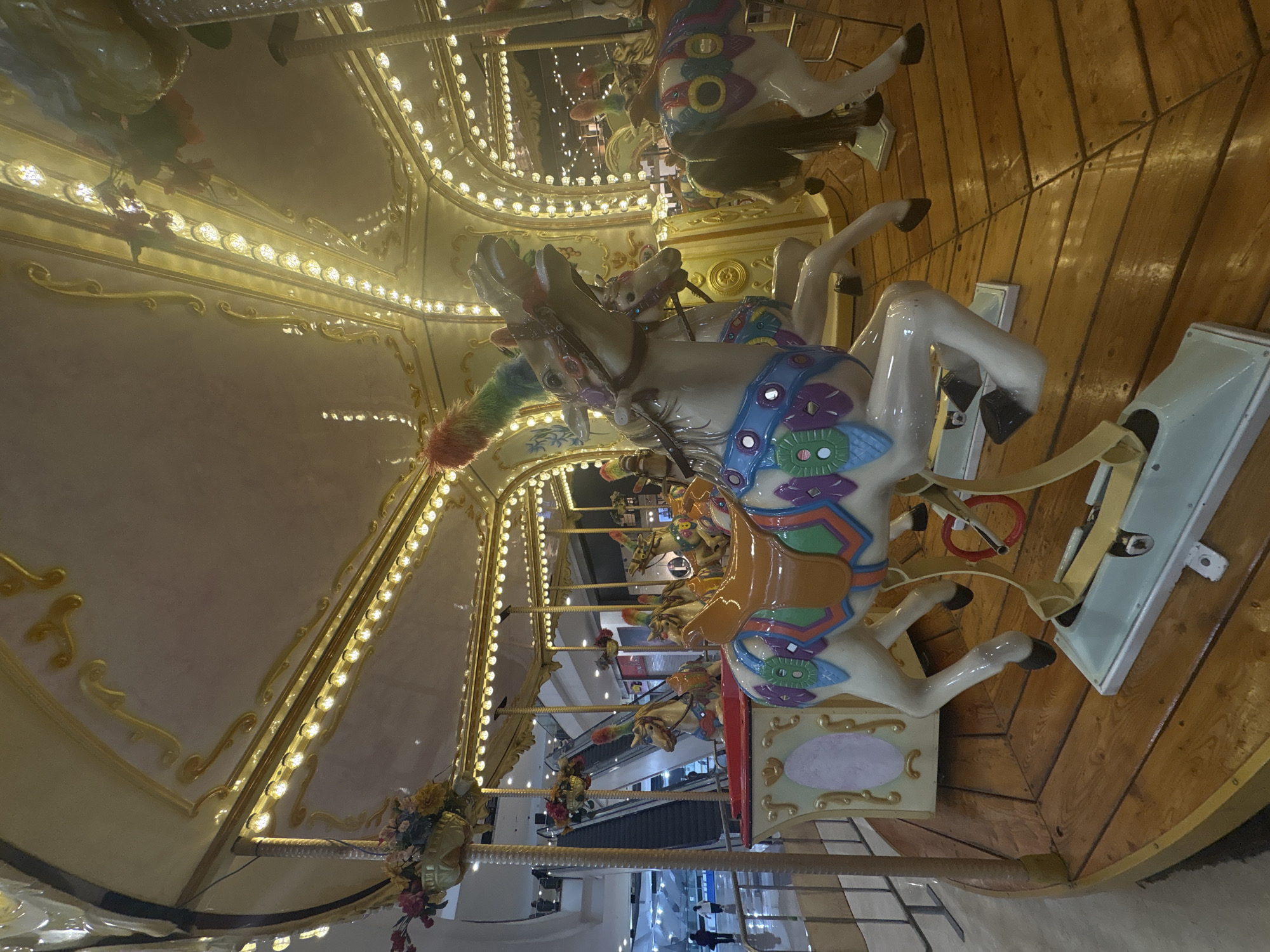 This image, rotated 90 degrees clockwise, captures an elaborately decorated carousel, likely situated indoors within a commercial or entertainment complex in Mexico City, Mexico. The perspective is from inside the carousel, looking across its ornate structure and into the background.
The central subjects are several carousel horses, predominantly white with vibrant, multi-colored saddles, bridles, and decorative patterns. Some horses feature fluffy, rainbow-colored tails, adding to their whimsical appearance. These horses are stationary, positioned on a polished wooden platform that makes up the carousel's floor. Each horse has a pale blue-green base or footrest.
Above the horses, a decorative, cream-colored canopy or ceiling portion of the carousel is brightly illuminated by numerous small, round, yellow lights arranged in rows and within golden, scrolling patterns. Additional embellishments include artificial flower arrangements in shades of red, pink, and yellow, attached to the carousel's support poles.
In the background, beyond the immediate carousel structure, a multi-level indoor environment is visible. Distant artificial lights illuminate what appears to be escalators and other architectural features of a shopping mall or large public space. A few blurred, indistinct human figures can be seen in the far background, indicating the presence of other people in the area. The overall scene exudes a warm and festive atmosphere, characteristic of indoor entertainment venues, particularly at what appears to be evening or night due to the reliance on artificial lighting. No discernible text is visible on the carousel itself or in the immediate surroundings.