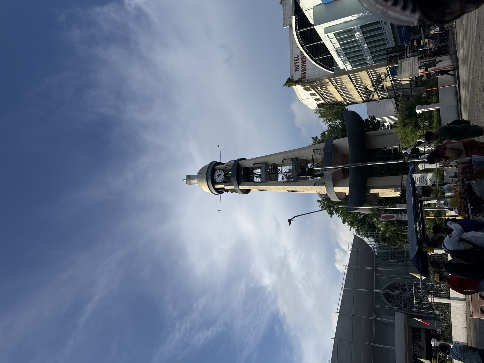 The image shows a low-angle view of the Magsaysay Park Clock Tower in Davao City, Philippines. The tower, a prominent landmark, is made of concrete and has a circular base with external spiral stairs. A clock face is visible near the top of the tower. Adjacent to the tower is a multi-story building with the signage "HOTEL MAGUINDANAO" in red lettering. To the left of the tower, a large building with scaffolding suggests ongoing construction or renovation. In the foreground, several individuals are seen near a street vendor's cart, which is laden with food items and drinks. The sky is a mix of blue with scattered white clouds. The scene depicts a typical urban environment with elements of local landmarks, businesses, and daily life.