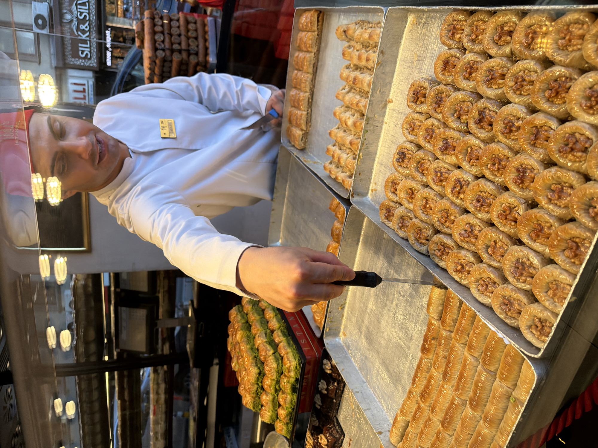A man wearing a white uniform and a red fez is standing behind a display of baklava at a food establishment in Istanbul, Turkey. He is holding a knife and appears to be cutting a piece of baklava. The display case contains various types of baklava, some with pistachios and others with walnuts. In the background, there are shelves filled with more baked goods and signage for "Silk & Silver." The lighting suggests an indoor setting, likely a bakery or dessert shop.