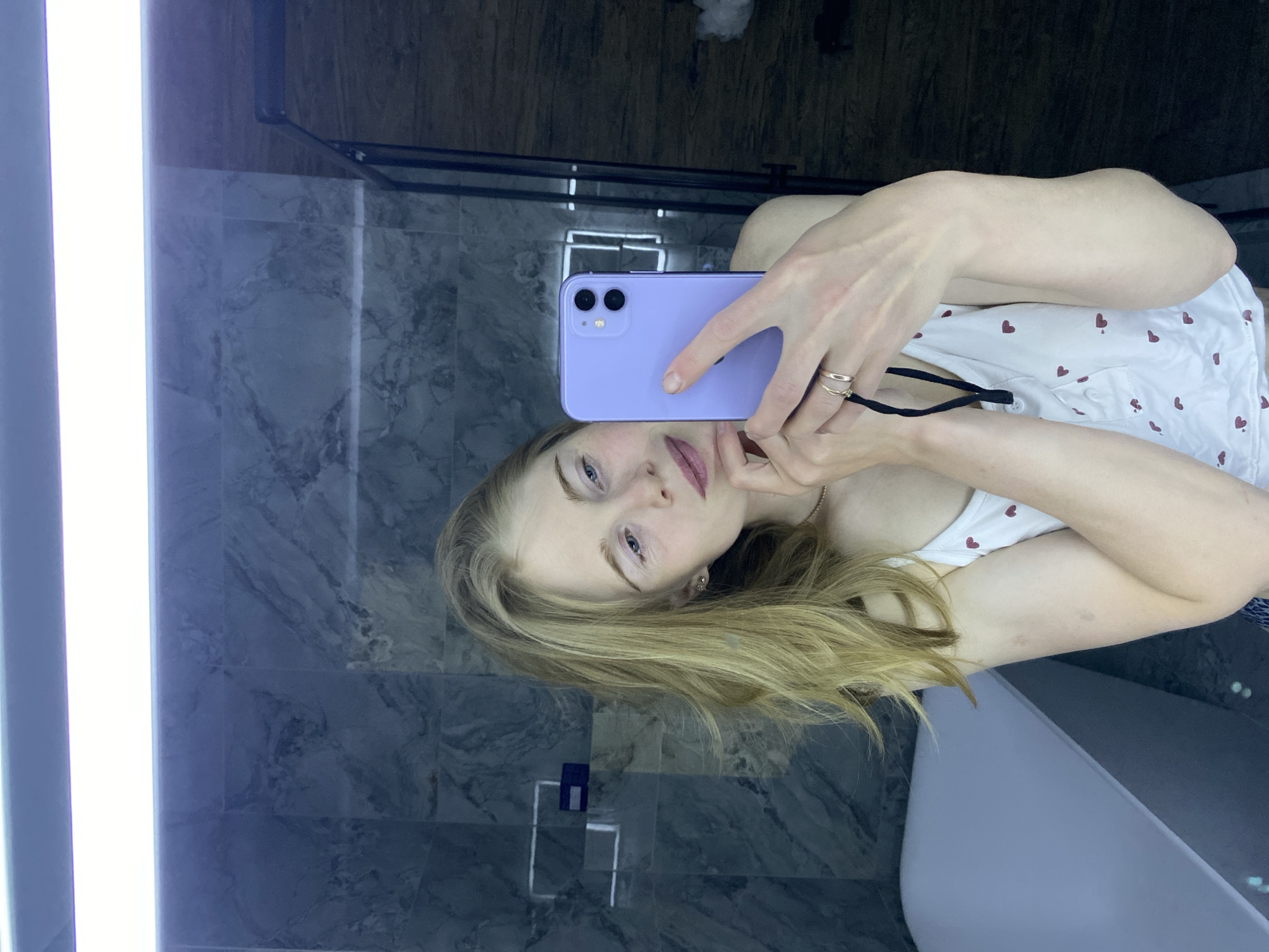 A woman is taking a selfie in a bathroom. She is holding a purple smartphone in her right hand, and her face is partially obscured by the phone. She has long, blonde hair and blue eyes. She is wearing a white tank top with red hearts. The bathroom has gray marble walls and a white bathtub. There is a bright light fixture on the left side of the frame and a chrome towel rack in the background. The location is Chaikovsky, Russia.