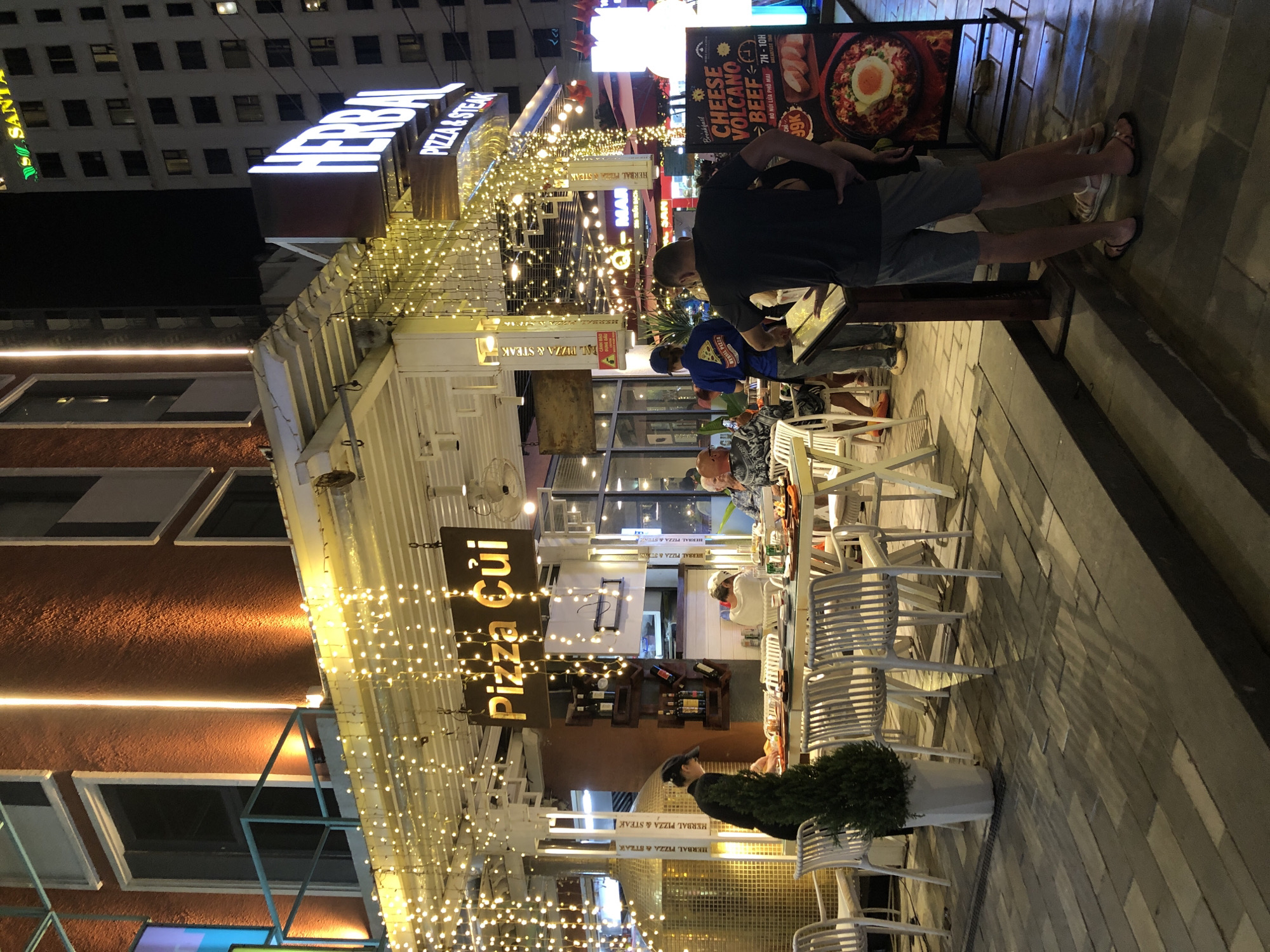 The image captures an exterior view of a restaurant in Da Nang, Vietnam, at night. The restaurant, named "Pizza Cùi," is adorned with numerous strings of small, warm-toned fairy lights. The building's facade features orange textured walls with vertical white strip lights. In the foreground, a person wearing a black cap and uniform is seen arranging items on a table, suggesting they are a server. Several white outdoor dining chairs and tables are set up on a paved walkway. A few patrons are seated at the tables, and others are standing, including a man in a dark t-shirt and shorts looking at a menu board. To the right, a large sign displays food items, including "Cheese Volcano Beef." The general ambiance is that of a lively, open-air dining establishment.
