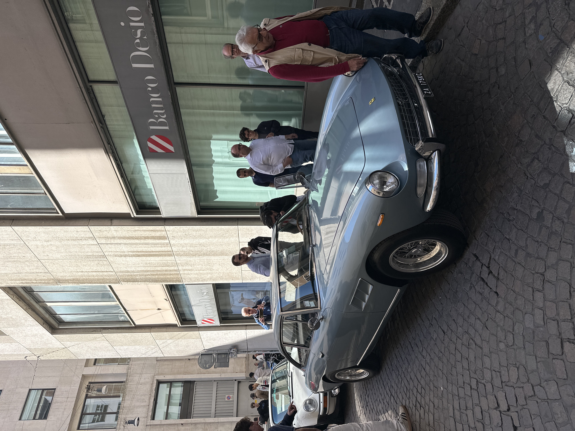 A light blue classic car is parked on a cobblestone street in Milan, Italy, in front of a building displaying the “Banco Desio” sign. Several people are standing around and observing the car. Two men are leaning over the hood of the car, examining it. Other individuals are positioned behind them, and a few seem to be taking pictures. The street is partially shaded, and the overall setting gives the impression of an urban environment. A white car is partially visible behind the blue car. The Italian license plate "ZA 175KC" is visible on the front of the blue car.
