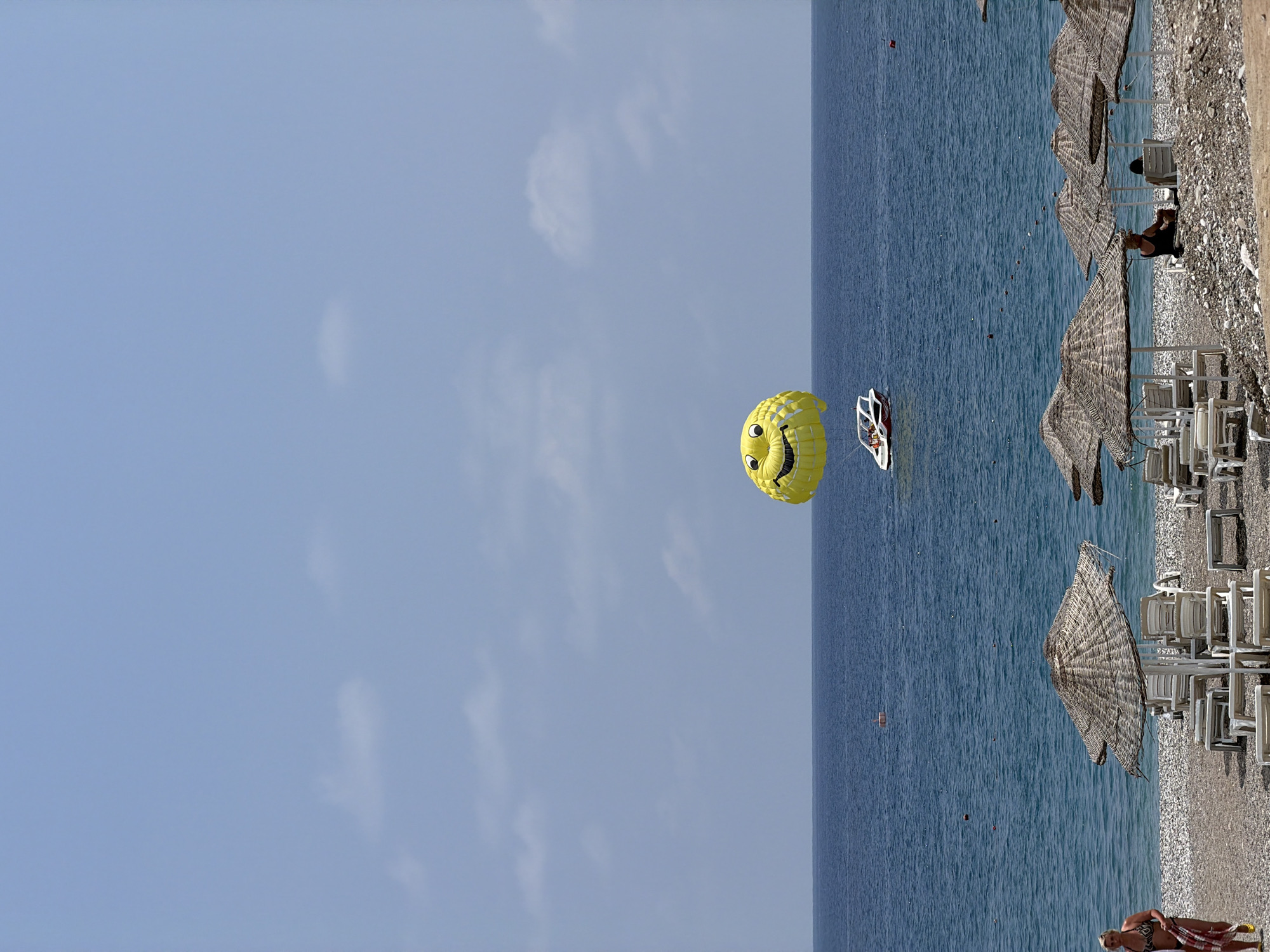 The image shows a beach scene with the ocean in the background. The sky is blue with scattered clouds. In the water, a parasail in the shape of a smiley face is being towed by a boat. Several beach umbrellas and lounge chairs are visible on the pebbled beach. A woman in a bikini is walking on the beach, holding a towel. Two people are sitting under a beach umbrella. The location is listed as Unknown, Unknown.