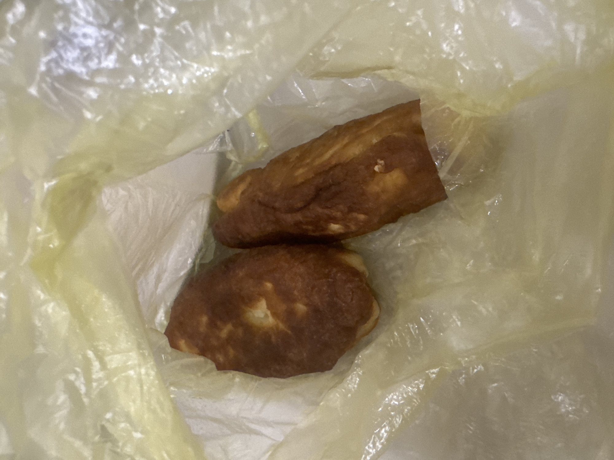 Two fried pastries are visible within a translucent yellow plastic bag. The pastries are golden brown in color and appear to be of irregular shapes. The bag is creased and somewhat crumpled, suggesting it is a disposable grocery bag. The background is indistinct and appears to be a plain surface, possibly a table or counter. The image was taken in Saratov, Russia.
