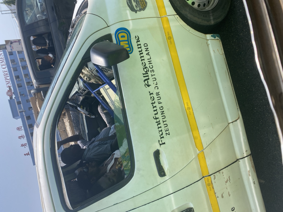 The image depicts a daytime scene in an urban environment, likely in traffic, within the city of Unknown, Ghana.
A white van or taxi occupies the left and bottom portion of the frame. The vehicle shows signs of wear, including paint chips and scratches on its lower body and door. It features horizontal yellow reflective tape with "DVLA" markings. A prominent black decal on the side reads "Frankfurter Allgemeine ZEITUNG FÜR DEUTSCHLAND," accompanied by a circular blue and yellow "MD" logo.
Inside the white van, three dark-skinned males are visible through the open driver's side window. The driver, wearing a light-colored collared shirt, is seated with his left arm raised, looking towards his right. The front passenger, wearing sunglasses and a white sleeveless top with a colorful pattern, is looking towards the camera. A third passenger, also in a white sleeveless top, is partially visible behind the front passenger, appearing to be resting.
To the right of the white van, a dark-colored pickup truck is partially visible. A person wearing a white face mask is seen through its open rear passenger window, looking towards the camera.
In the background, a multi-story blue building stands under a clear sky. This building displays prominent red signage, including Chinese characters "王者集团" (King's Group) and "EMPIRE CEMENT" with smaller "AN" and "FAN" text. An area of disturbed earth or construction material is visible between the vehicles and the background building. The vehicles appear to be stationary or moving slowly.