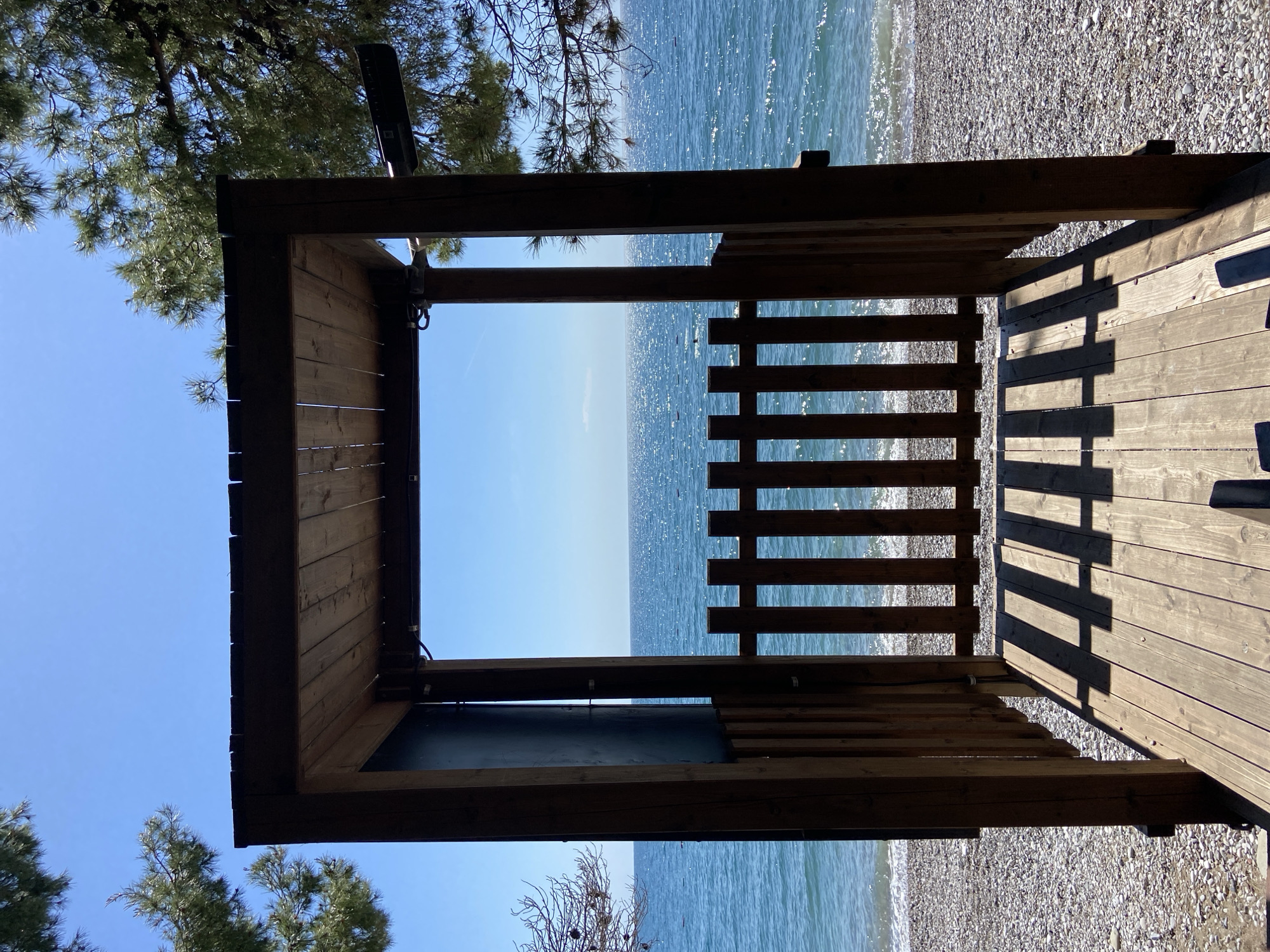 The image presents a serene coastal scene viewed from within a rustic wooden structure. The wooden frame of a shelter or pergola occupies the foreground, with a sturdy wooden deck forming the floor and a roof above. On the right side, a slatted wooden railing or fence extends towards the sea. Sharp, distinct shadows from the structure and railing are cast across the wooden deck, indicating bright, direct sunlight, suggesting it is mid-morning or early afternoon.
Beyond the wooden structure, a narrow pebble beach meets the sparkling, blue-green waters of the sea. The sea stretches calmly to the horizon under a clear, azure sky. The water's surface glistens with countless reflections of the sun. Pine trees with green needles and visible trunks are seen in the upper left and lower right (when rotated correctly) of the frame, suggesting the structure is nestled amidst a natural, shaded environment. A long, dark light fixture is mounted on the underside of the wooden beam on the left side of the structure.
No people or visible text are present. The setting evokes a peaceful and inviting atmosphere, typical of a sunny day at a Mediterranean beach, consistent with the location context of Bar, Montenegro.