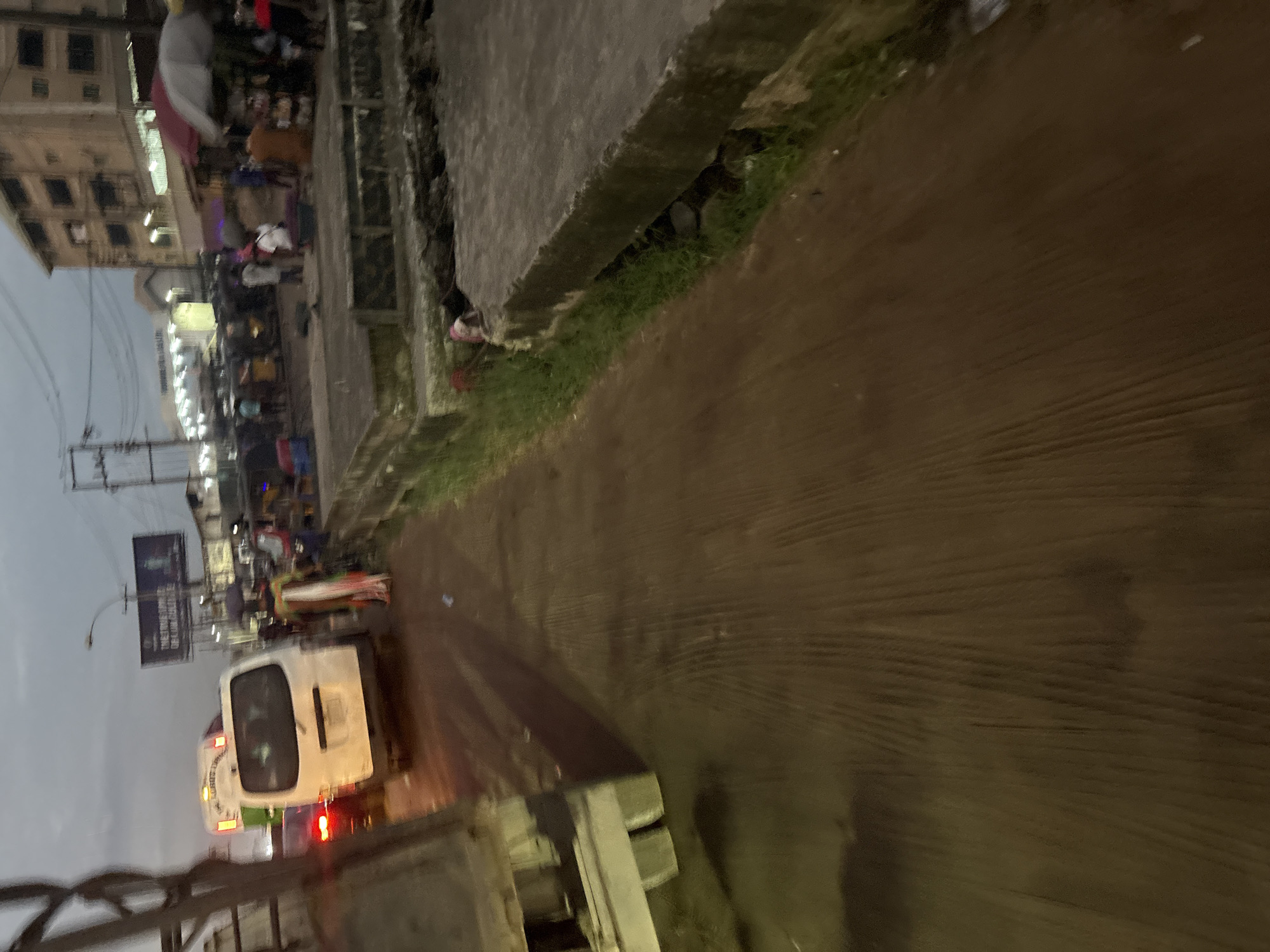 This image captures a busy street scene in Onitsha, Nigeria, likely during dusk or overcast conditions. The foreground is dominated by a reddish-brown, textured surface, possibly a road or pavement, with visible tire tracks and some debris. To the left, the side of a white truck is visible.
In the midground, several vehicles are present, including a white passenger van and a red vehicle behind it. The van has a license plate that is partially obscured but appears to be yellow. A person in colorful traditional attire is walking near the van, holding what looks like a dark umbrella. Further back, a market area is visible with stalls and people. Buildings with multiple stories form the backdrop, some appearing weathered. A large billboard with text is mounted on a pole.
The scene depicts a typical urban environment in Onitsha, characterized by traffic, commercial activity, and a mix of modern and older infrastructure. The presence of market stalls and diverse vehicles suggests a lively and active part of the city. The lighting indicates either early morning or late afternoon.