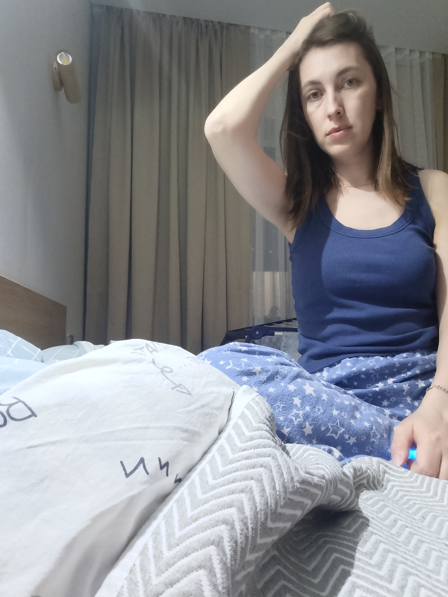 An adult female, appearing to be in a bedroom in the city of Chaikovsky, Russia, is depicted seated on a bed, looking directly at the camera. She has brown hair and wears a dark blue ribbed tank top and light blue pajama pants patterned with white stars. Her left hand is raised, with fingers resting on the top of her head. Her right hand, wearing a thin chain bracelet, exhibits a faint blue light emanating from an unseen object. The bed features a light wooden headboard, a light blue patterned sheet, a white pillow with dark grey symbols and text, and a grey and white chevron-patterned blanket. A cylindrical wall-mounted light fixture is visible on the upper left wall. Grey curtains cover a window in the background, with a partially visible clothes drying rack positioned behind them.
