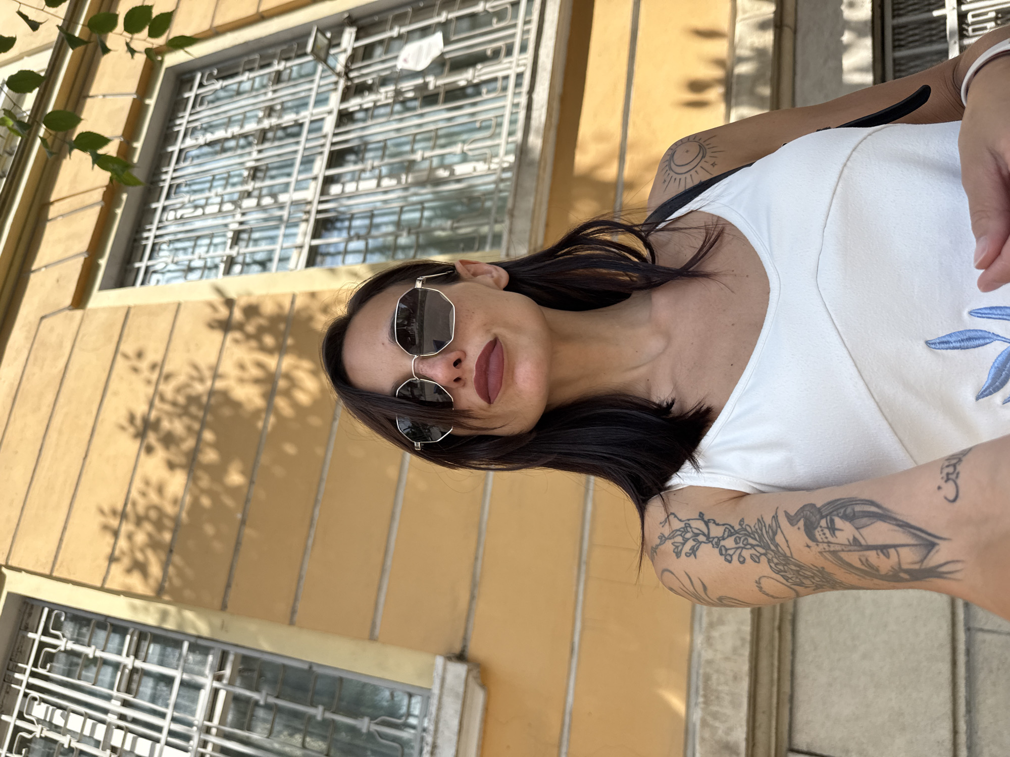 In Pleven, Bulgaria, a woman stands in front of a yellow building with barred windows. She wears a white tank top and sunglasses, and has tattoos on her arms, one depicting a face and another a sun and moon. The sun casts dappled shadows on the building's facade.