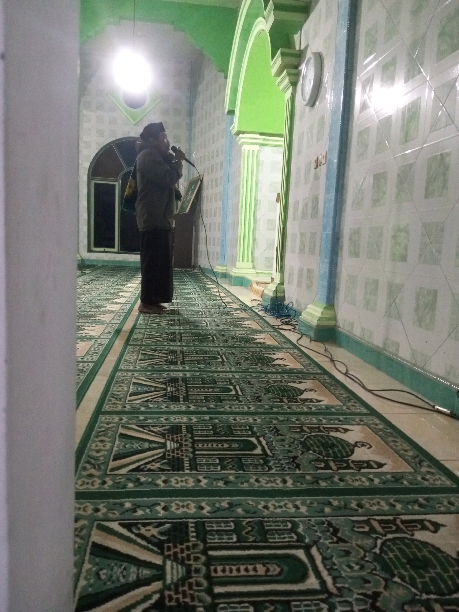 A man stands in a hallway of a mosque, holding a microphone and speaking into it. He is wearing a kufi, a traditional cap, a jacket, and sarong pants. The hallway is carpeted with green and beige prayer mats that have intricate designs. The walls are tiled with a light green and white pattern, and there are light green pillars supporting arched doorways. A clock is visible on the wall. The lighting in the mosque is dim, with a bright light source overhead. The scene appears to be within the city of Andongsili, Indonesia.