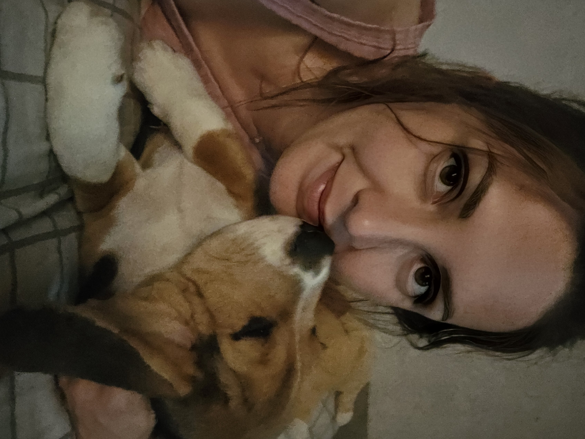 A woman is lying on her side, likely in bed due to the plaid bedding visible, and is close to a beagle puppy. The puppy, predominantly brown and white, is nestled against her chest. The woman has dark hair and brown eyes. She is smiling slightly. The location is Unknown, Unknown.