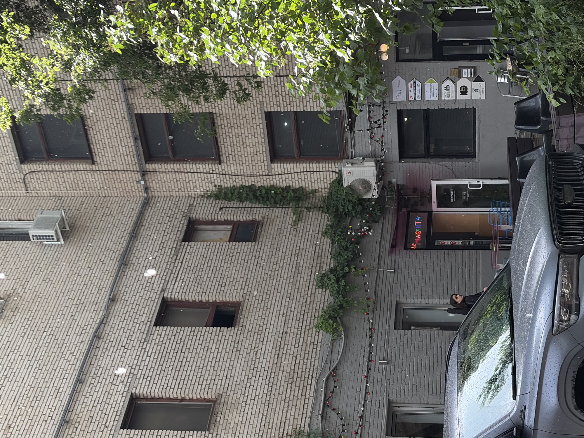 The image shows the exterior of a building with light-colored brickwork. Several windows are visible, some with dark-tinted glass. An air conditioning unit is mounted on the exterior wall. Green foliage hangs from a tree in the upper left corner, casting shadows. Strings of small lights, some red and white, are draped along the side of the building. A person is partially visible leaning out of a window on the right side of the frame, looking to their right. A silver vehicle, possibly a van, is parked in front of the building, with its windshield reflecting the surrounding greenery. The overall scene suggests an outdoor urban environment. The time of day appears to be daytime, with sunlight filtering through the leaves. The presence of the vehicle and the architectural style suggest an urban setting, potentially a street or courtyard. No specific event or activity is clearly depicted, other than the presence of the person in the window and the parked vehicle. There is some visible text on signs or plaques on the wall to the right, including "Pret" and what appears to be "LE MAGENTA".