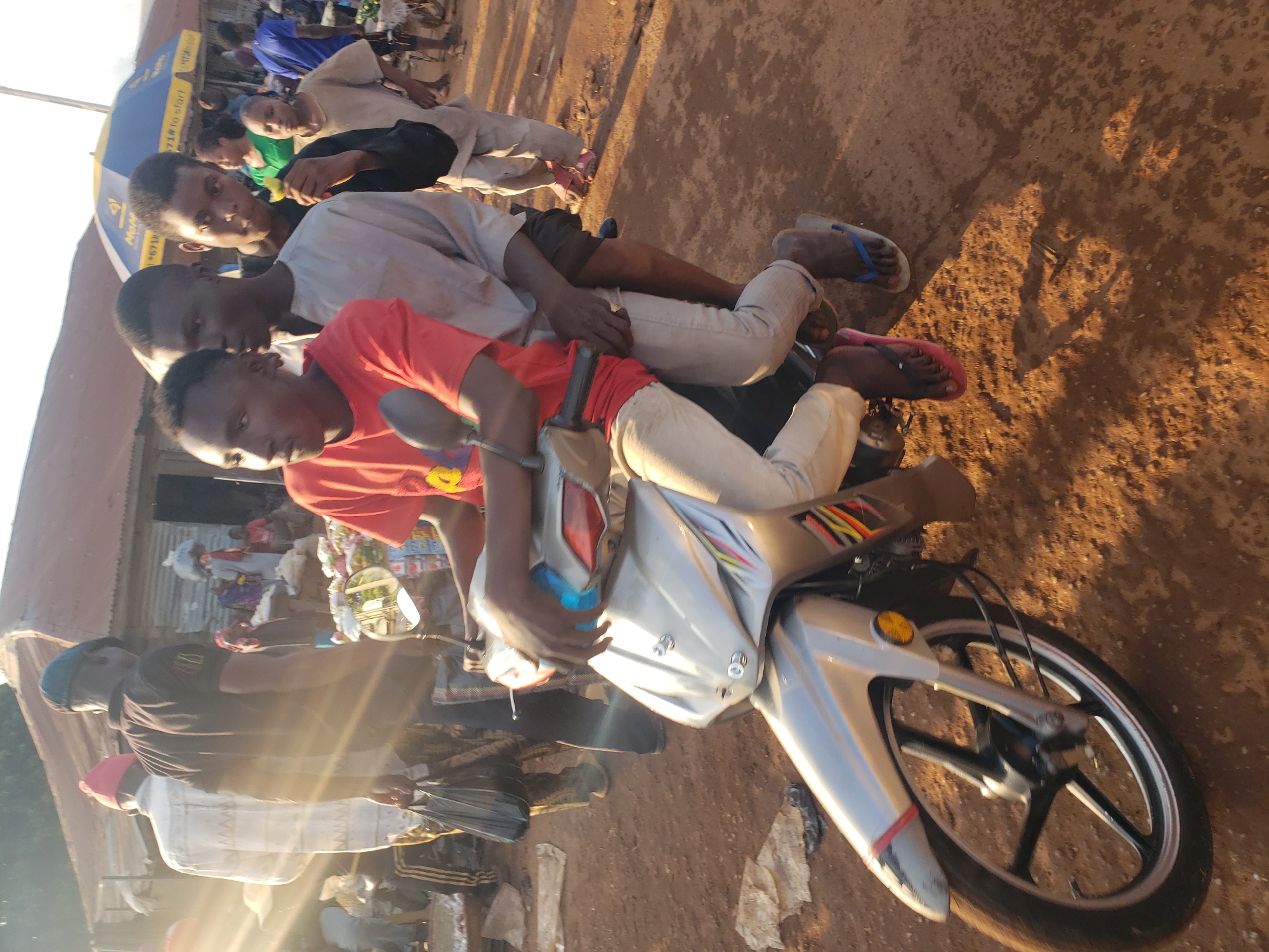 In Kakuri Gwari, Nigeria, a street scene unfolds with a group of young men on a motorcycle. The rider, in a red t-shirt, is followed by two others, one in a light-colored shirt and another behind him. They are on a silver motorcycle with visible branding, possibly indicating its model or origin. The environment is dusty, with a dirt road surface illuminated by the sun, casting shadows. In the background, there are structures that appear to be market stalls or informal shops with corrugated metal roofing. Other individuals are visible in the background, some carrying loads and others engaged in commerce. A blue and yellow umbrella is present, suggesting a marketplace setting. The overall impression is one of a bustling, everyday scene in a Nigerian town, with a focus on the young men and their mode of transportation.