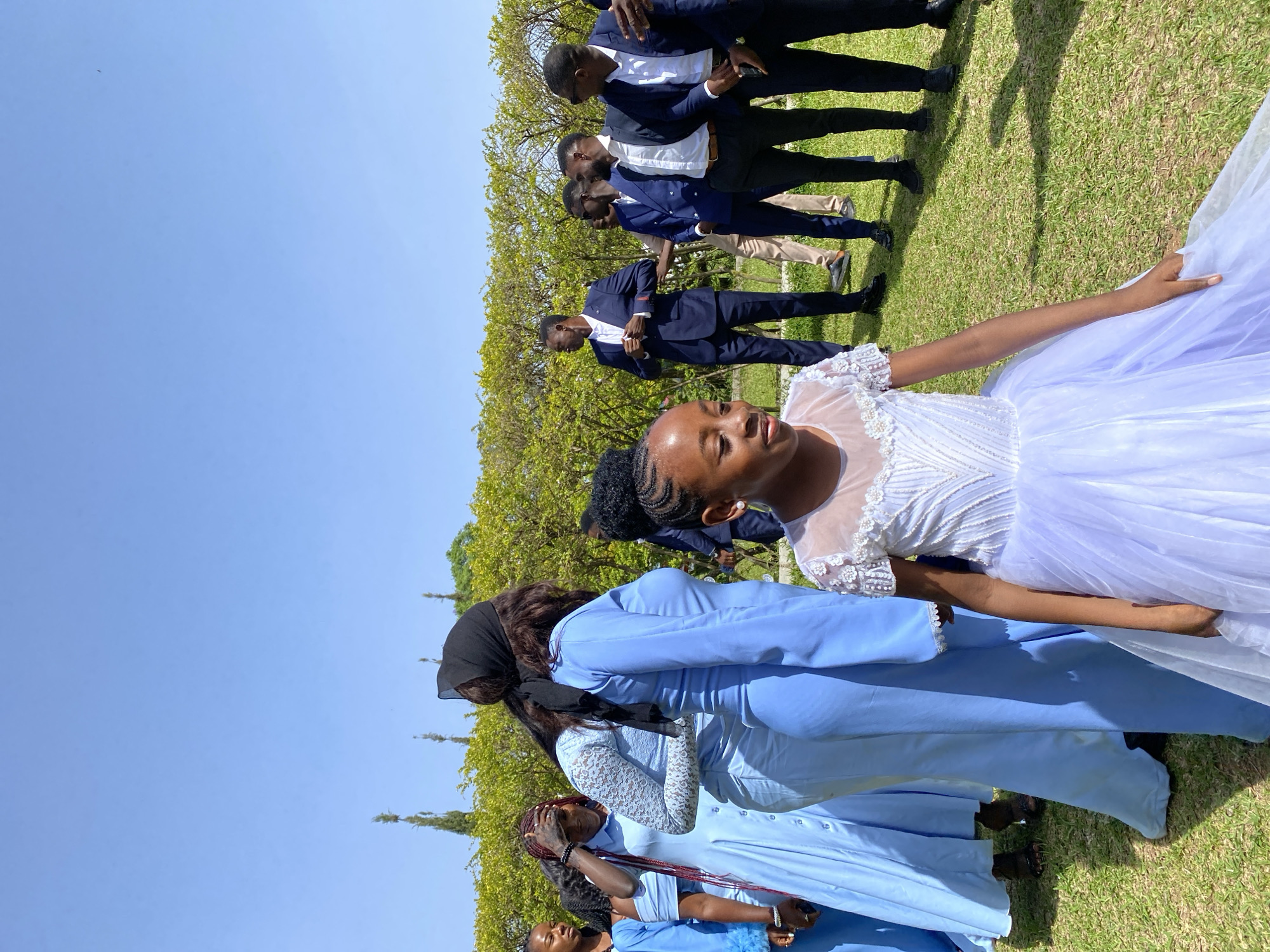 The image shows a group of people outdoors in Bukuru, Nigeria. A young girl in a white dress stands out in the foreground. Behind her are several women dressed in light blue outfits. To the left of the women, some men are dressed in suits and are walking on the green grass. The sky is blue, and there are trees in the background.