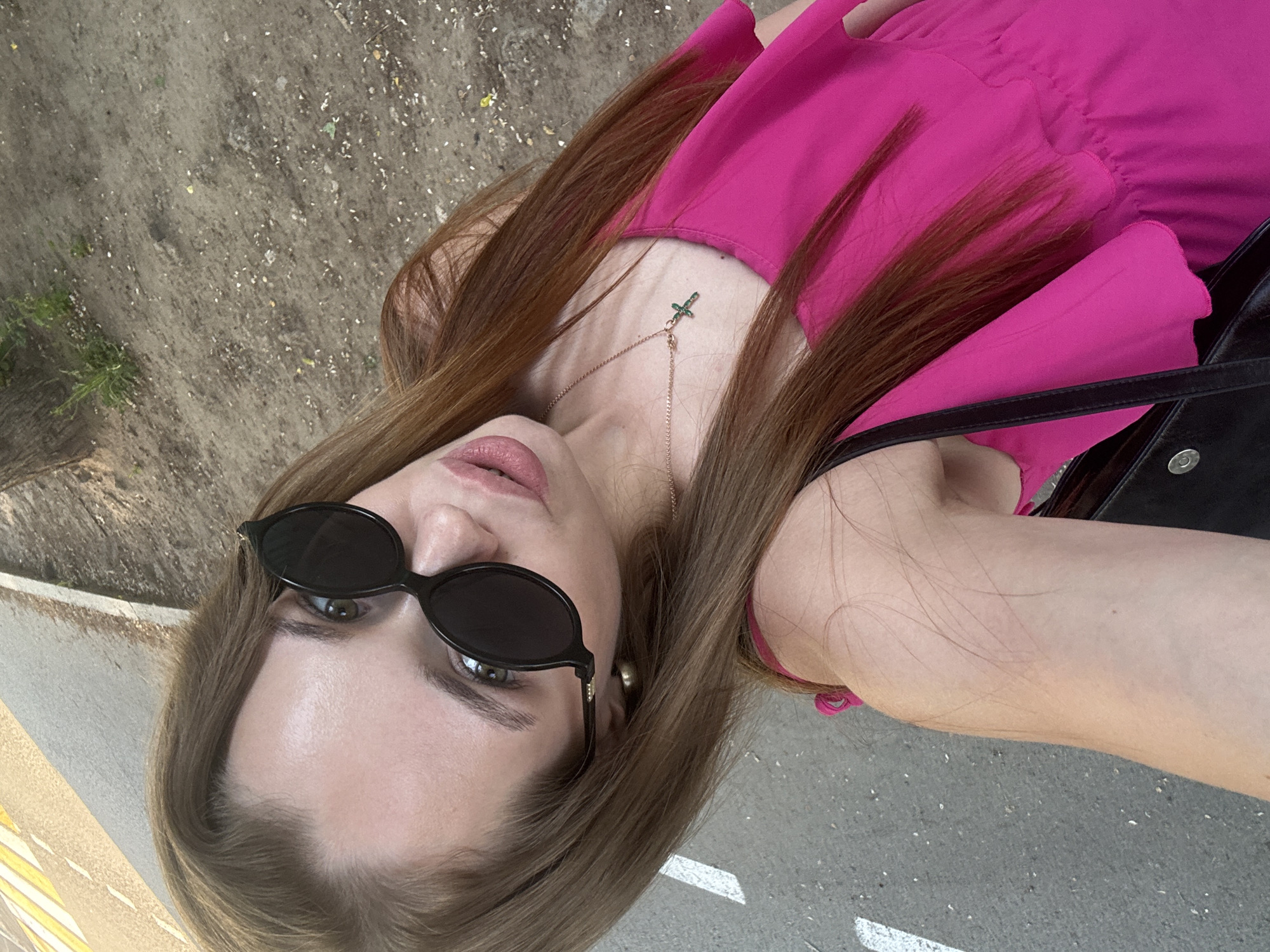 A woman is seen from a top-down perspective, wearing sunglasses and a bright pink dress with a gold necklace. She is outdoors on a paved surface, possibly a street or sidewalk, in the city of Chaikovsky, Russia. The surface has white lane markings and appears to be adjacent to an area with grass and some sparse vegetation. The woman has shoulder-length brown hair and is holding what appears to be a black purse or bag.
