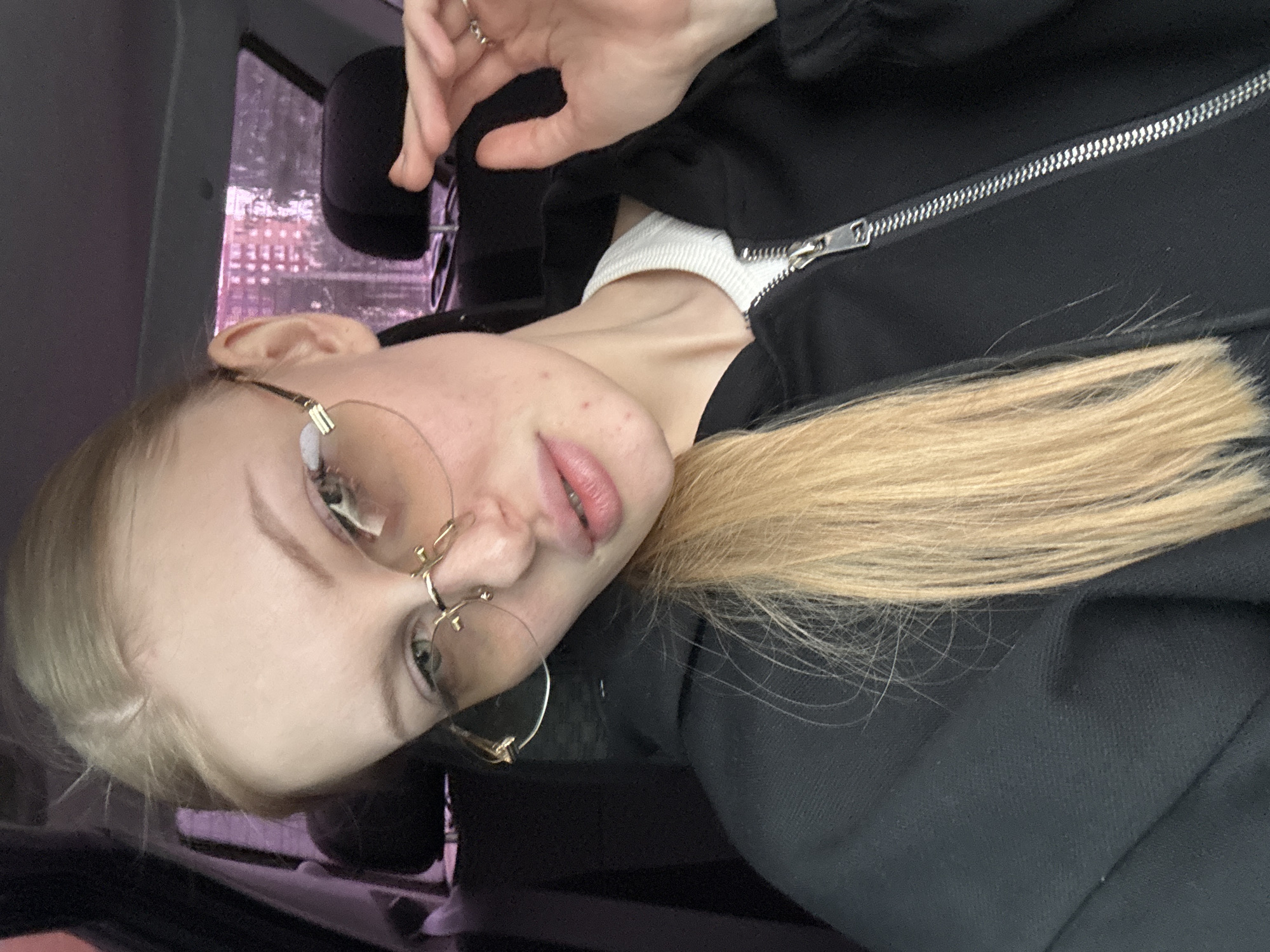 A blonde-haired woman wearing glasses and a black jacket is photographed from a close-up, canted angle. The location appears to be inside a vehicle, possibly a car, given the window and interior elements visible in the background. Through the window, there are indistinct buildings suggestive of an urban environment. Based on visual cues, the location is presumed to be in Perm, Russia. The woman has light skin and is wearing glasses. Her hair is styled to one side. She is wearing a black jacket with a visible zipper. Her expression is neutral. Her left hand is raised near her head. The lighting is natural.