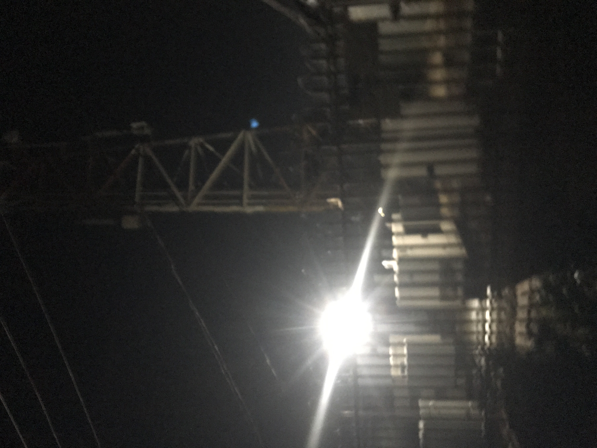 The image depicts a nighttime scene at a telecommunications tower located in Rafin Par, Nigeria. A bright light source, possibly a security light or a camera flash, causes significant glare and lens flare, obscuring details in the foreground. Behind the glare, a tall, lattice-style tower, likely a telecommunications or radio tower, is visible. The tower is constructed of metal beams forming a triangular structure. A fence, constructed of vertical metal posts and horizontal bars, runs across the foreground, partially obscured by the bright light. The background is dark, with the sky appearing black, suggesting the absence of significant ambient light. Some faint electrical wires are visible in the upper left portion of the image. The overall impression is of an industrial or functional area at night, dominated by the illuminated tower and the artificial light.