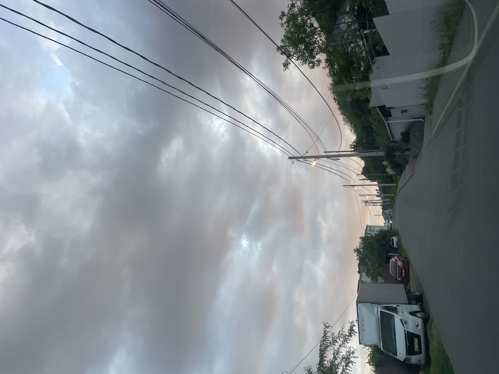 The image depicts a downward sloping street in Baranovka, Russia, under a cloudy sky with hints of pink and orange hues. The street is paved and appears to be residential, with houses and fences lining both sides. On the left side of the road, a white cargo van is parked, followed by a red car and a white sedan further down the street. Overhead, power lines are strung across the sky, supported by utility poles. Green trees and vegetation are visible throughout the scene, suggesting a somewhat rural or suburban setting. The overall atmosphere is overcast, possibly indicating the approach of evening or a change in weather. There are no people visible in the image.