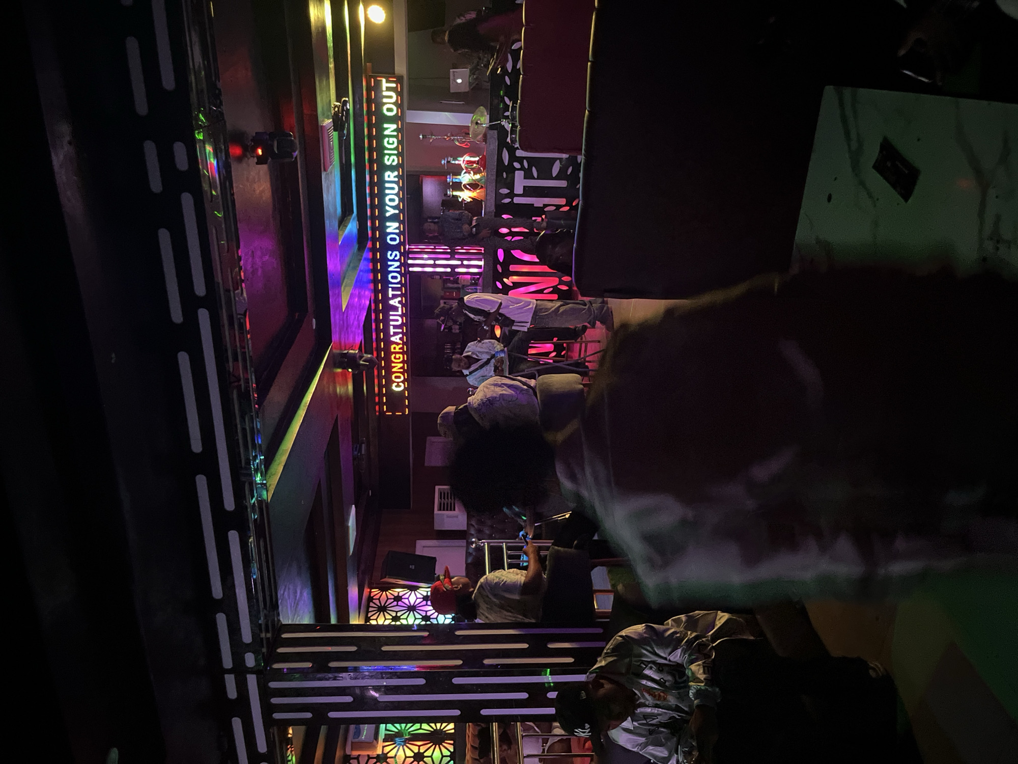 The image depicts an indoor scene, likely a bar or club, illuminated by colorful neon lights and a large digital display board. The digital board displays the text "CONGRATULATIONS ON YOUR SIGN OUT". Several individuals are present, some seated, others standing. In the foreground, a person is seated with their back to the camera, partially obscured by shadow. To the left, another individual in a light-colored jacket and a baseball cap is seated. Further back, near a bar area, several people are standing and interacting. The bar is equipped with bottles and what appear to be hookah pipes. The environment features decorative pillars with vertical light strips and patterned glass panels. The overall atmosphere suggests a social gathering or celebration.