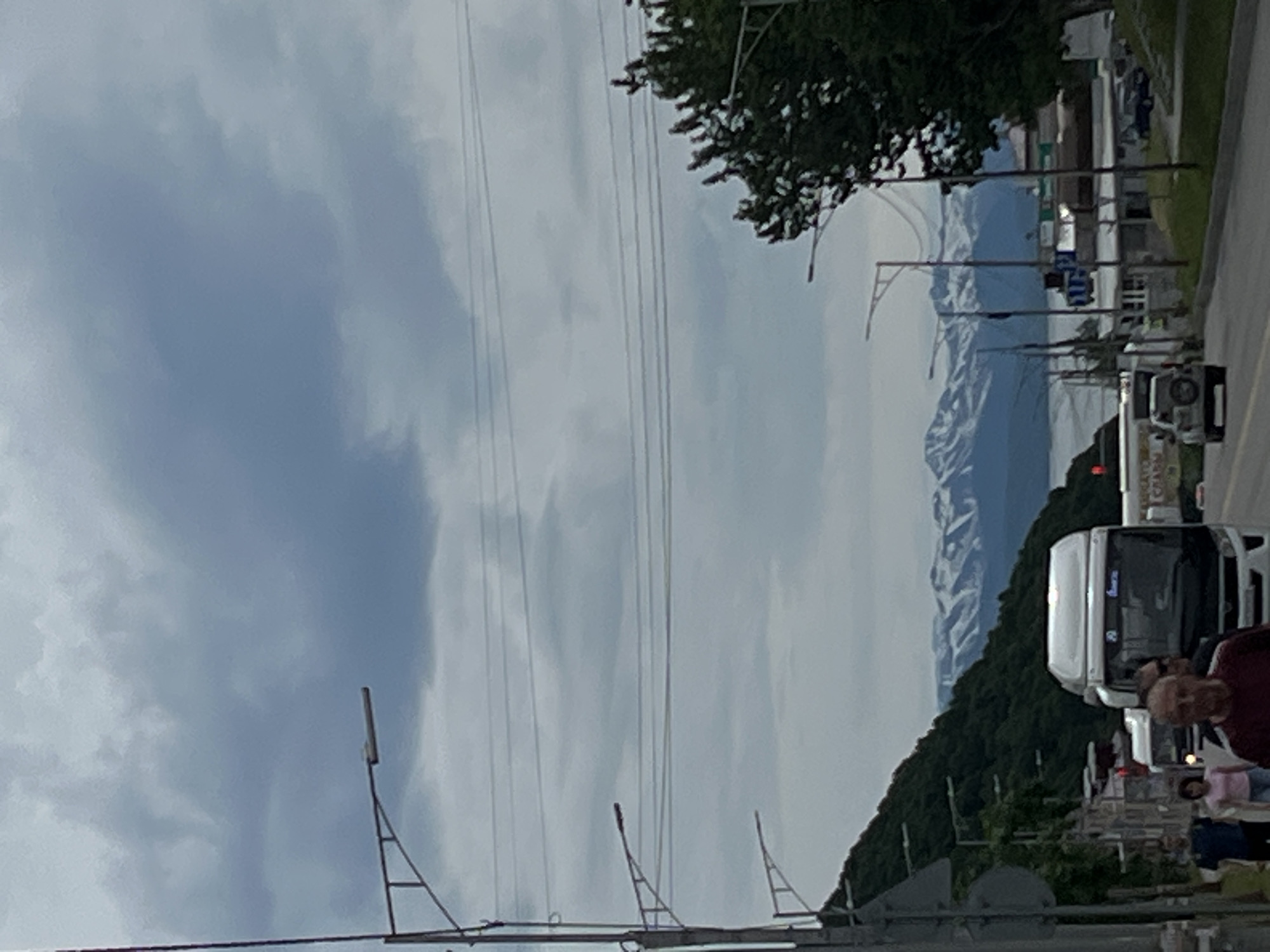 The image depicts a view of a street in Petropavlovsk-Kamchatsky, Russia, with a backdrop of snow-capped mountains and a body of water, likely the Avacha Bay. The sky is overcast with heavy gray clouds. A road runs through the scene, with several vehicles present. On the right side of the road, there are buildings and a lush green tree. On the left side, a steep, tree-covered hill slopes upwards. Several streetlights are visible along the road. In the foreground, there are pedestrians walking on the sidewalk. A white bus is prominent in the middle of the road, and a silver SUV is further down the street. The overall impression is one of a cloudy day in a scenic coastal city.