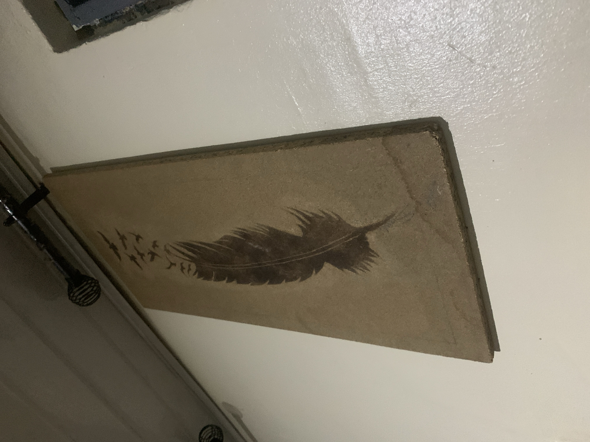 A vertical, rectangular piece of pressed particle board, approximately 2.5 feet long and 1 foot wide, is mounted on a textured, off-white wall. The particle board has a textured, light brown surface, resembling unfinished wood or cardboard. It displays a dark brown silhouette of a feather with intricate barbs, positioned vertically and centered on the board. Above the feather, a flock of small, dark birds in silhouette appears to be ascending or scattering upwards from the feather's quill. The board is affixed to the wall with its top edge slightly angled upwards from left to right. Above the artwork, a dark, thin pipe runs horizontally across the ceiling, with two black, cage-like light fixtures suspended from it. The left light fixture is partially visible, and the right fixture is in full view. The wall has a slightly uneven, granular texture. To the right of the artwork, a dark, recessed opening, possibly a window or niche, is visible with rough, dark edges. The lighting in the scene is dim, casting subtle shadows.