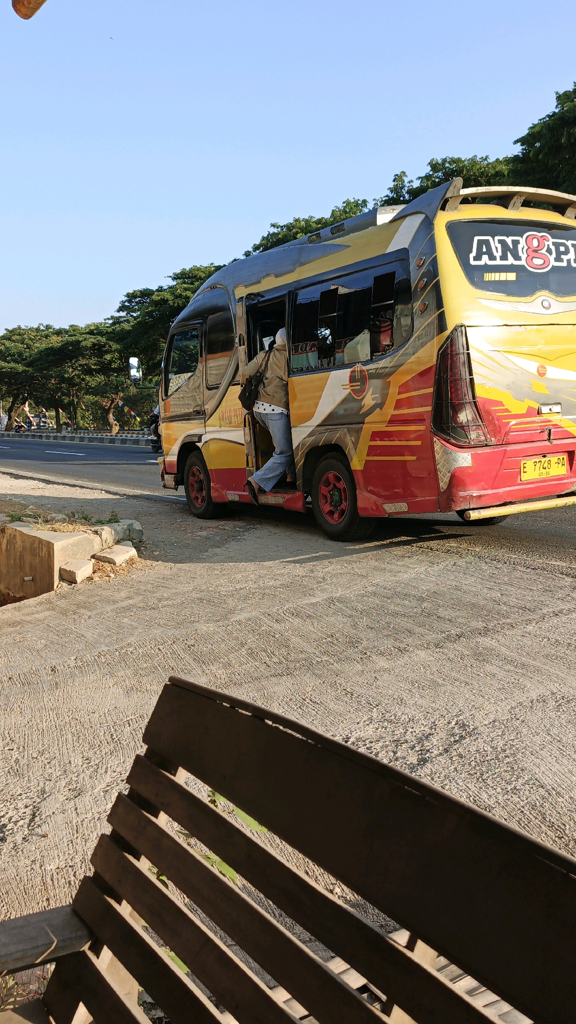 A yellow and red bus with the word "ANG&PI" written on its back is parked on the side of a road in Krimun, Indonesia. A person wearing jeans and a light-colored jacket is stepping out of the bus. The bus is decorated with colorful stripes and a logo on its side. In the foreground, a wooden bench is partially visible. The background shows a row of trees and a clear blue sky. The road appears to be paved, and there is a concrete ditch next to it. The scene suggests a stop along a bus route, possibly in a semi-urban or rural area of Krimun.