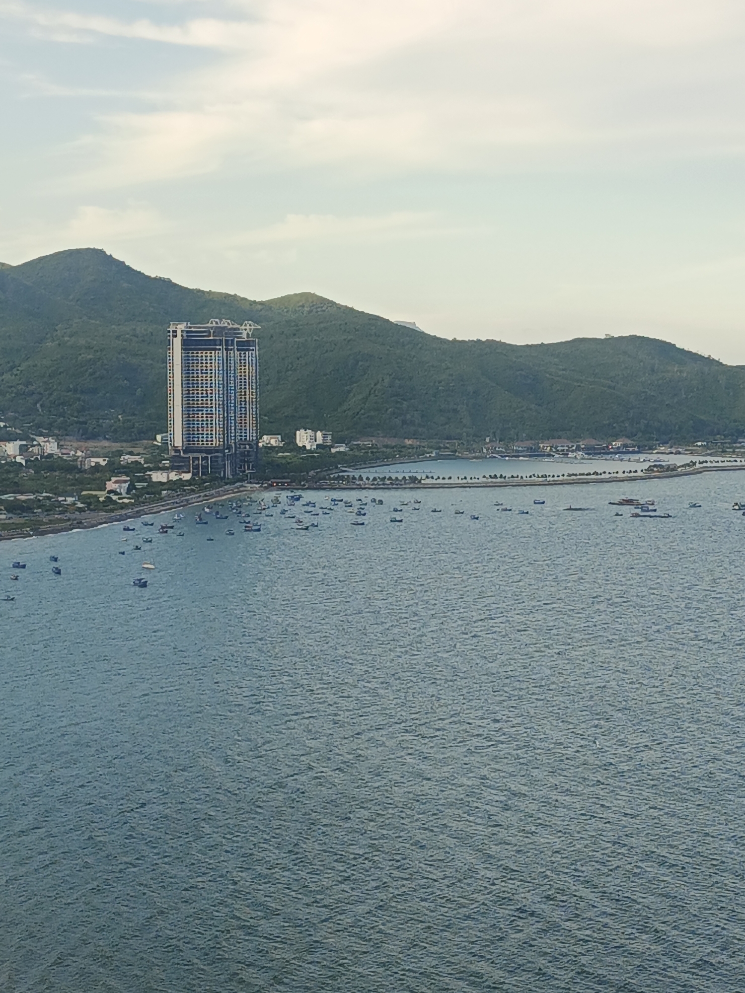 This is an aerial view of a coastal city, likely in Vietnam, showing a bay filled with numerous small boats. Dominating the left side of the frame is a tall, modern skyscraper with a distinct checkered facade of yellow and blue squares. Behind the skyscraper and the coastline, a range of lush, green mountains forms the backdrop. The sky is overcast with light clouds. The water is a tranquil blue, with small waves indicating a gentle breeze. The scene suggests a developing urban area with a strong connection to the sea.