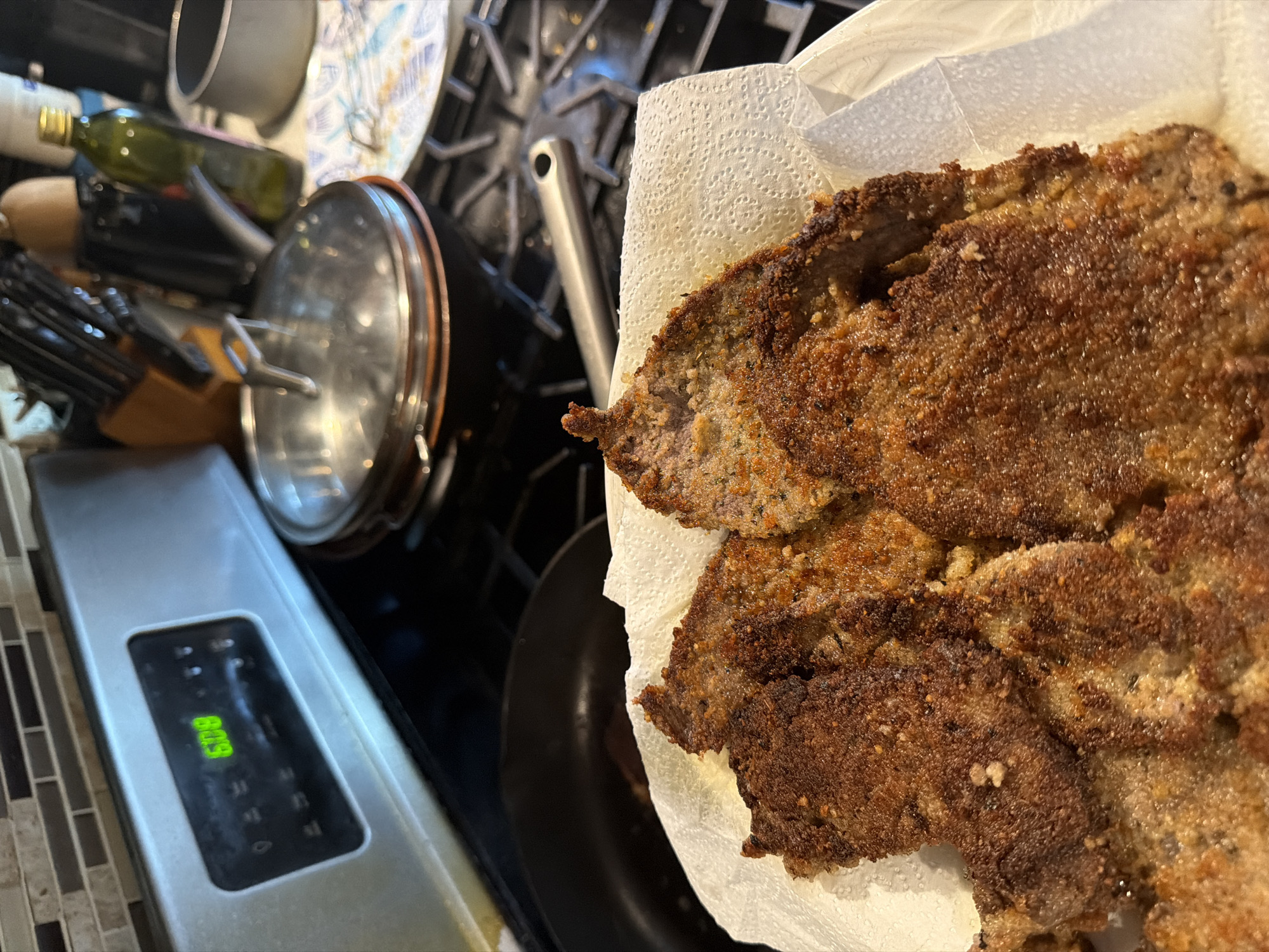 The image captures a close-up, slightly overhead view of a home kitchen scene, focusing on several freshly cooked, breaded cutlets resting on white paper towels. The cutlets are golden-brown and appear crispy, suggesting they have just been fried and are draining excess oil.
In the background, a gas stovetop with dark grates and a silver-colored pot or pan is visible, indicating recent or ongoing cooking activity. To the left, kitchen clutter includes a wooden knife block with several knives, a dark green bottle (likely olive oil), and a pot with a metallic lid. Further to the left, part of a kitchen appliance, possibly an oven or microwave panel, features a digital display showing the time "8:09" in green digits. The countertop has a mosaic tile backsplash composed of rectangular grey and dark grey tiles. The overall setting is a functional, lived-in kitchen. No people are visible in the image.