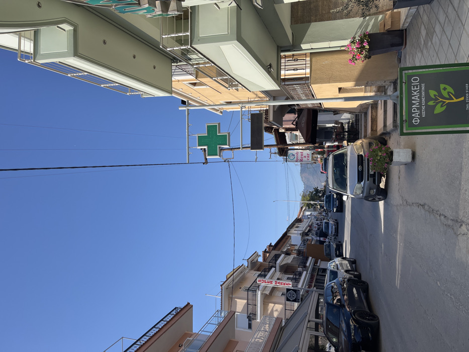 The image captures a street scene in Limenaria, Greece (latitude: 40.627, longitude: 24.575). The scene is dominated by a row of buildings lining both sides of the street, including a pharmacy with a green cross sign. The buildings appear to be of various heights and architectural styles. Several cars are parked on the street, some with their drivers visible behind the wheel. In the foreground, a silver SUV is parked in front of the pharmacy, with its front end facing the viewer. The street is paved and has a slight downward slope. There is a sign with a plant logo and the word "ΦΑΡΜΑΚΕΙΟ" (pharmacy) on a wooden stand in the lower right corner of the image. The sky is a clear blue, with a few wispy clouds visible. The overall atmosphere is calm and peaceful. There is a sense of normalcy and everyday life in the town.
