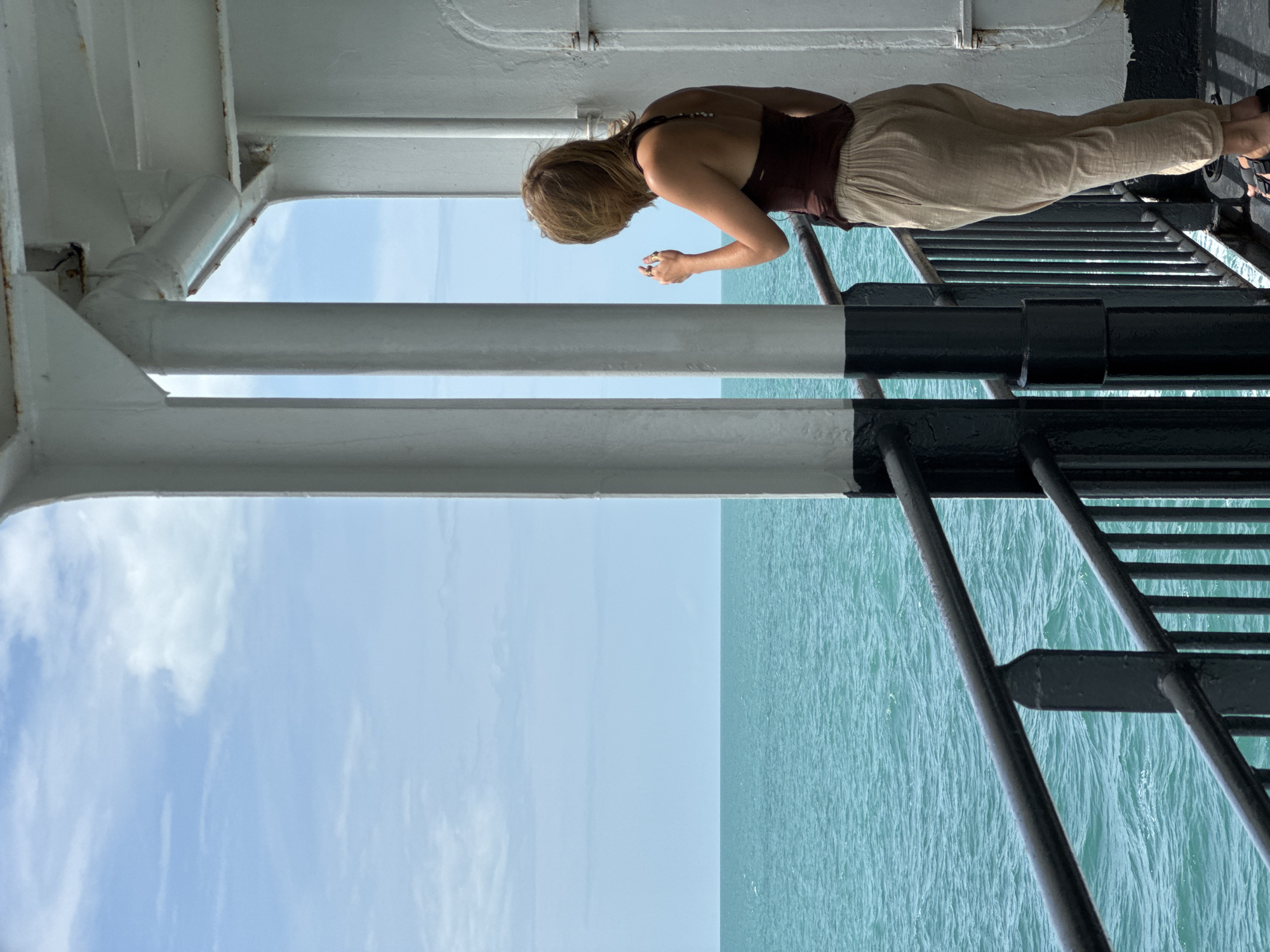 The image depicts a woman standing on the deck of a ferry boat, likely en route to or from Don Sak, Thailand. She is facing away from the camera, gazing out at the turquoise ocean and blue sky. The woman has shoulder-length blonde hair and is wearing a dark brown tank top and light-colored, loose-fitting pants. Her right hand is raised, holding what appears to be a phone or camera. The ferry's railing, painted black, is visible in the foreground, along with white structural supports and a portion of the boat's white exterior. The water is calm with slight ripples, and the sky is partially clouded.