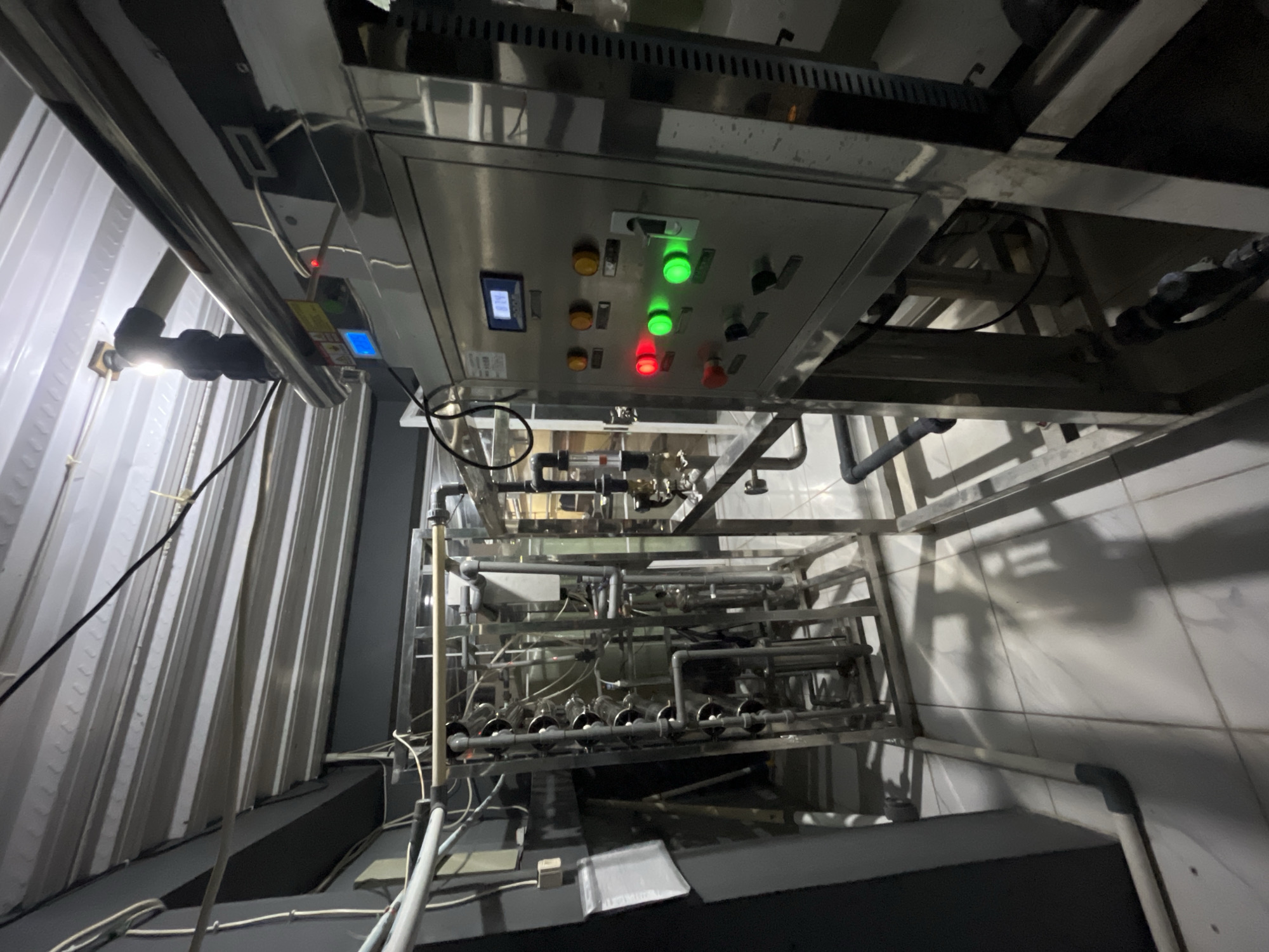 A water purification system is depicted in a dimly lit, industrial setting, likely a factory or processing plant. The machine features a stainless steel control panel with several illuminated indicator lights: two green lights are on, one red light is on, and three yellow lights are on. A digital display shows "819". Various pipes and tubes are connected to the system, forming a complex network. The environment appears to be a room with a corrugated metal ceiling and tiled flooring. Shadows are cast across the floor, suggesting a single light source. There are no people visible in the image. The specific location within Pekalongan, Indonesia, cannot be determined from the visual information provided.