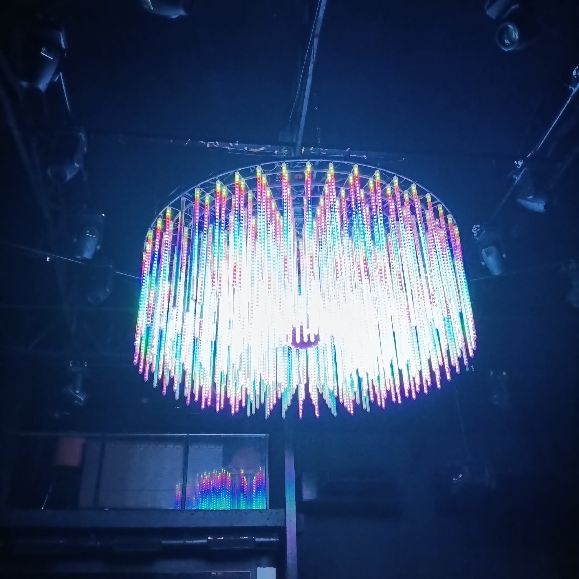 The image shows a large, illuminated chandelier-like structure in a dark environment, likely a club or event space in Sinquerim, India. The structure has a circular or semi-circular frame from which numerous illuminated rods or tubes hang down. The rods display a range of colors, including white, red, blue, green, and purple. The overall effect is a bright and colorful display against the dark background. The scene includes stage lighting fixtures mounted on the ceiling in the periphery of the chandelier. In the lower left corner, there's a visible shelf or platform. An indistinct human figure can be detected behind a transparent panel.