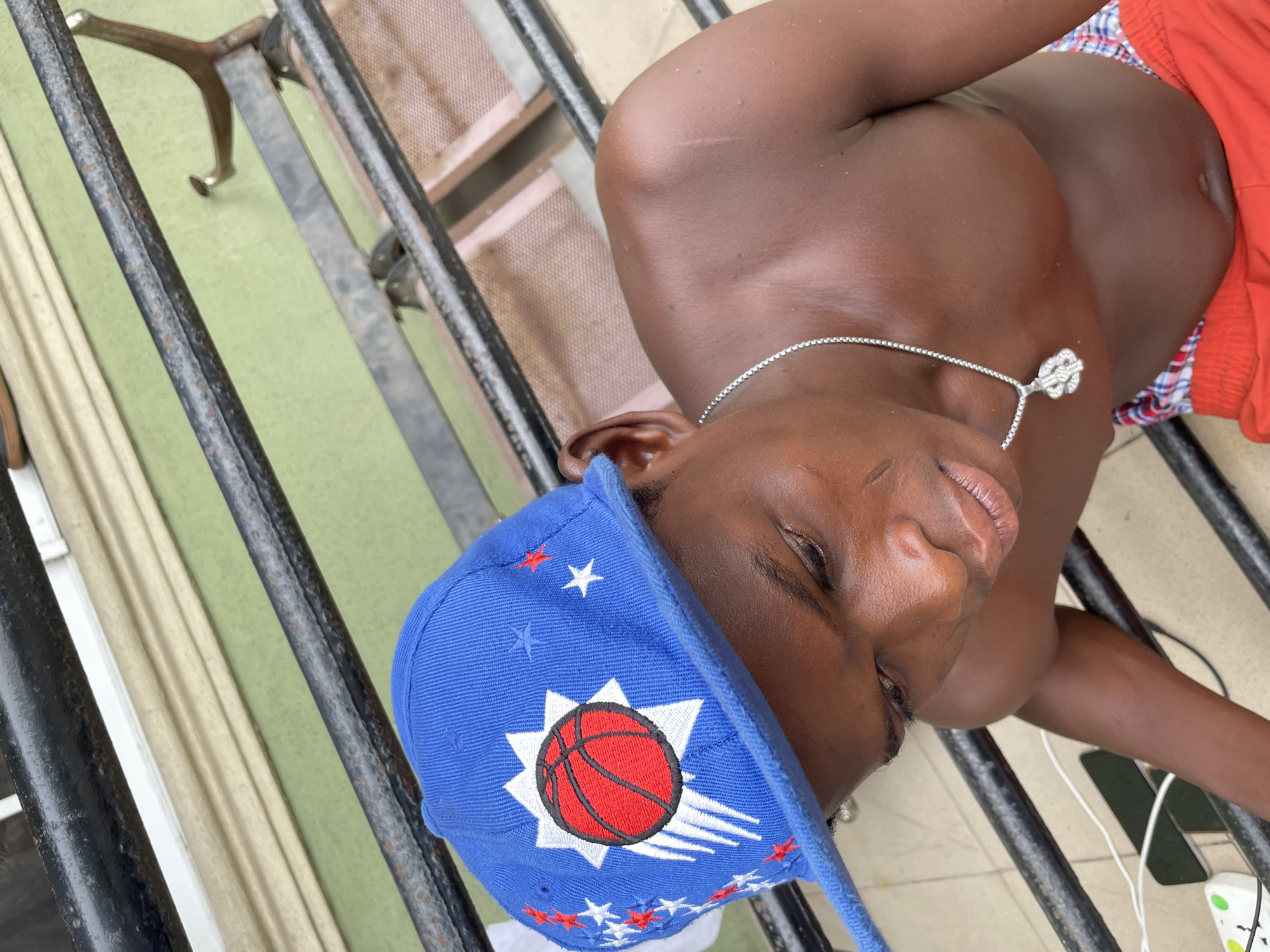 A medium-shot, eye-level photograph shows a dark-skinned person wearing a blue baseball cap with a red basketball design and white stars, lying sideways on a metal railing. The person is also wearing a silver chain necklace with a clover pendant. The person is in the city of Ado, Nigeria, lying on a metal railing. They are wearing red shorts and an object, likely a phone, appears to be attached to white earphones. The background features a green wall and a metal chair.