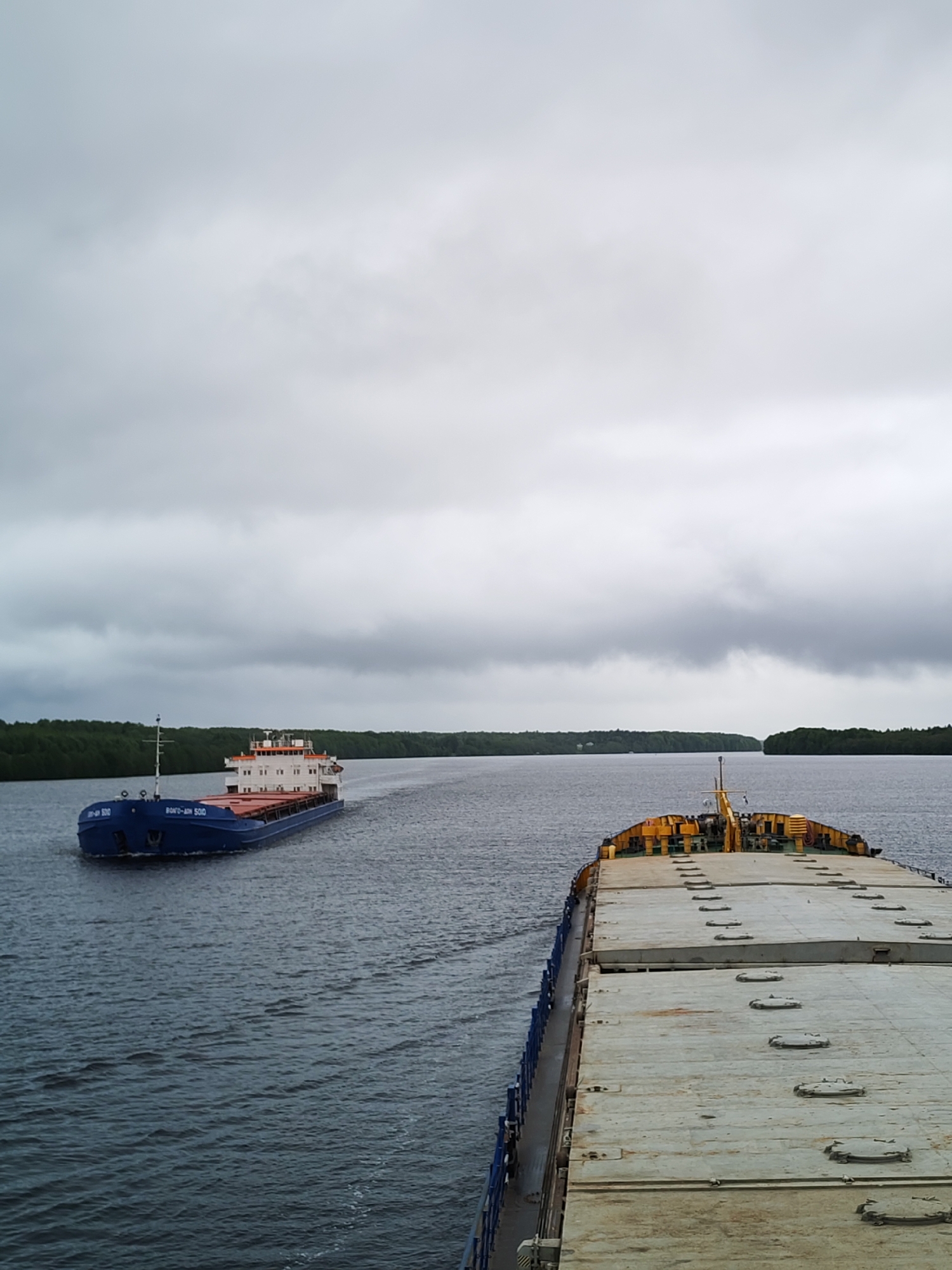 The image shows two barges on a wide river under a cloudy sky. Green tree lines the river's banks. One barge is blue and white, with lettering in Russian script. The other barge is in the foreground, showing a grey, flat deck with evenly spaced, circular hatches. A yellow structure sits at the barge's stern. The location is the city of Gorka, Russia.