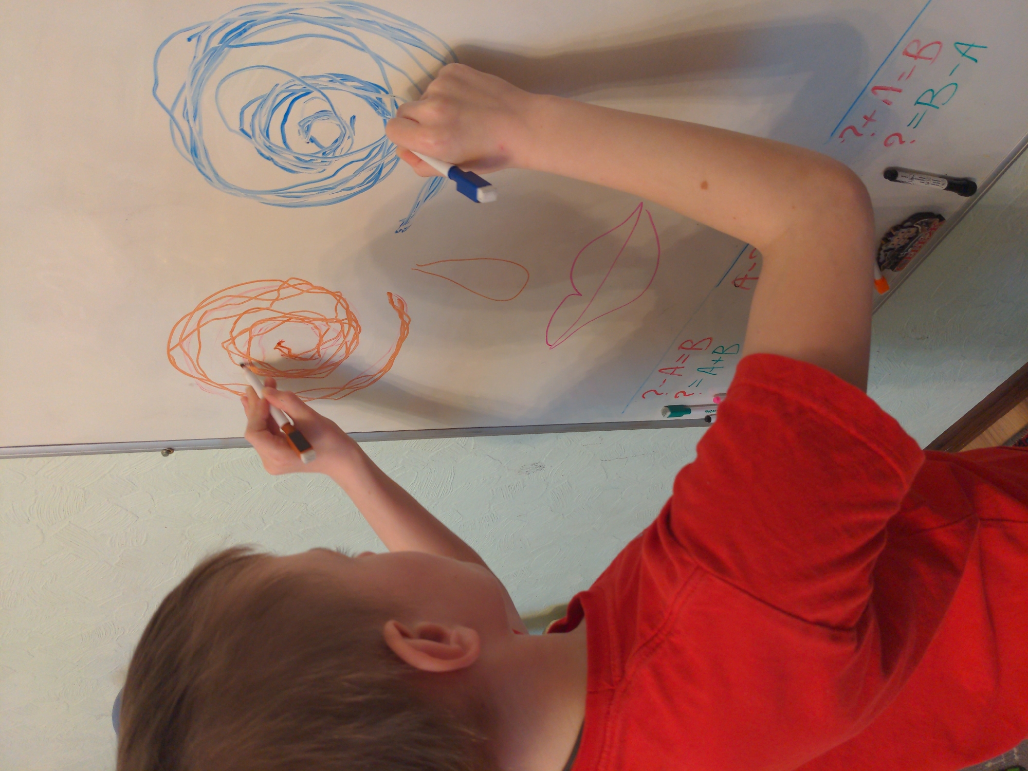 A child in a red shirt is drawing on a whiteboard. The whiteboard has multiple drawings, including a blue swirl at the top, an orange swirl below it, and two leaf shapes. Additionally, there are some equations written on the whiteboard, such as "?-A=B" and "?+11=B". The child is holding a marker in each hand and is actively drawing on the whiteboard.