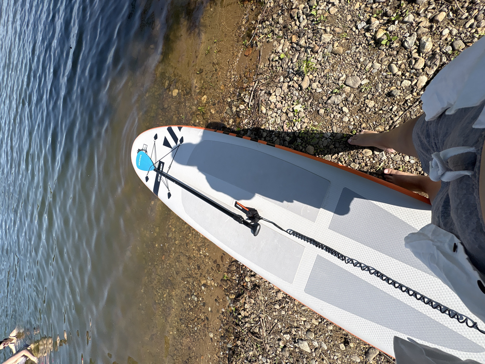 A first-person perspective shot shows a stand-up paddleboard resting on a rocky shore next to a body of water. The paddleboard is white and gray with orange accents and has a blue paddle attached. A black coiled leash connects the paddleboard to an ankle strap. Two people are partially visible in the water in the upper left of the frame. The water is clear near the shore, revealing the rocky bottom, and becomes darker further out. The sky is blue with some clouds.