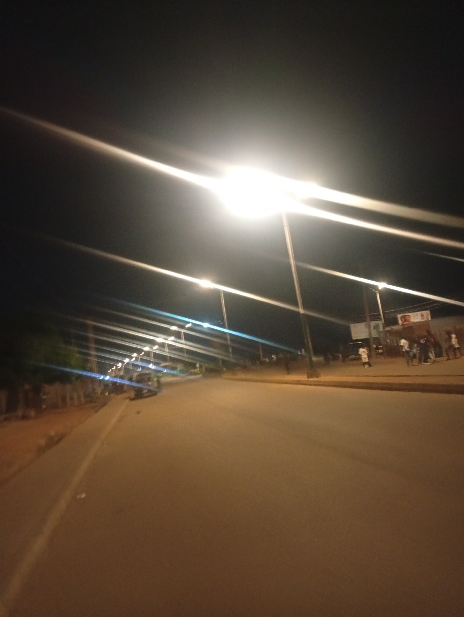 A nighttime street scene in Bukuru, Nigeria, is depicted. The image is dominated by the glow of streetlights, which create strong lens flares that streak across the dark sky. A paved road runs diagonally from the bottom right to the middle left of the frame. Along the left side of the road, a sidewalk is visible, bordered by a low fence and some trees. On the right side of the road, a few people are gathered near buildings. One building has a sign with a picture on it. Vehicles are present in the background, and one appears to be a small truck or van. The overall atmosphere is that of a quiet urban evening.