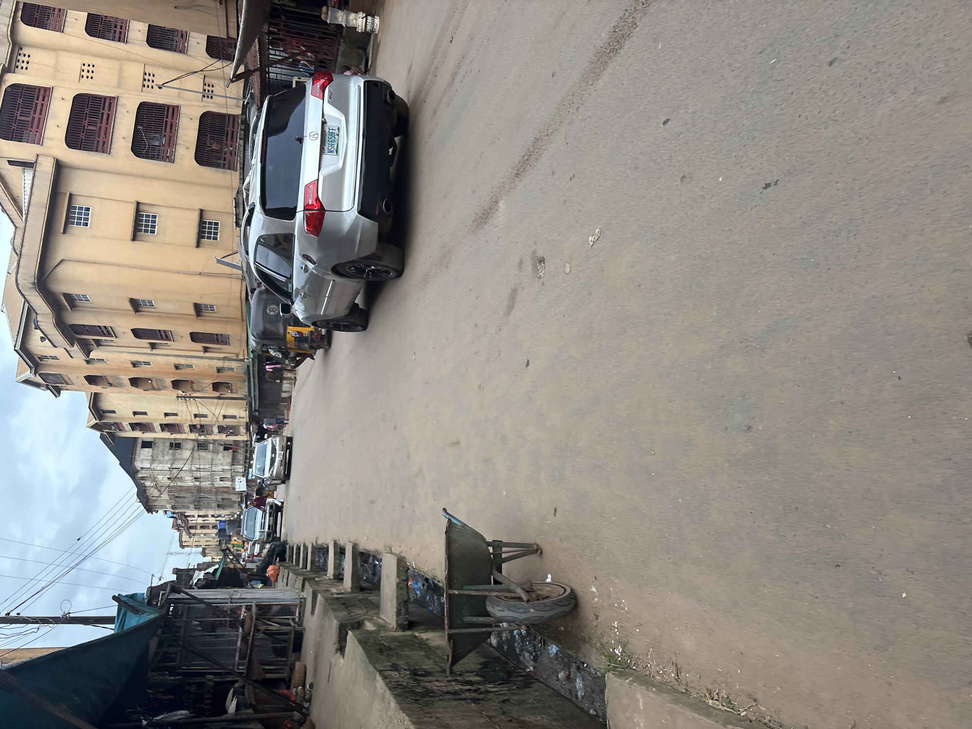 An exterior, medium shot captures a street scene in Onitsha, Nigeria, under an overcast sky. In the foreground, a weathered green wheelbarrow rests on the edge of a paved road, beside a concrete drainage channel filled with debris. The road, which appears to be unpaved in places, stretches into the distance. A silver SUV with a license plate reading "NSH 658FF" is the most prominent vehicle, seen from the rear as it drives away. Behind it, a three-wheeled auto-rickshaw, known as a 'Keke Napep', is also visible, with a person riding in the back. Further down the street, two other cars, a white SUV and a silver sedan, are moving. The sides of the road are lined with multi-story buildings, their facades painted in various shades of beige and yellow, with some exhibiting signs of wear. A market stall with a blue awning is set up on the left side of the road, displaying goods. The overall environment appears to be a bustling urban area with signs of commercial activity and typical street conditions.