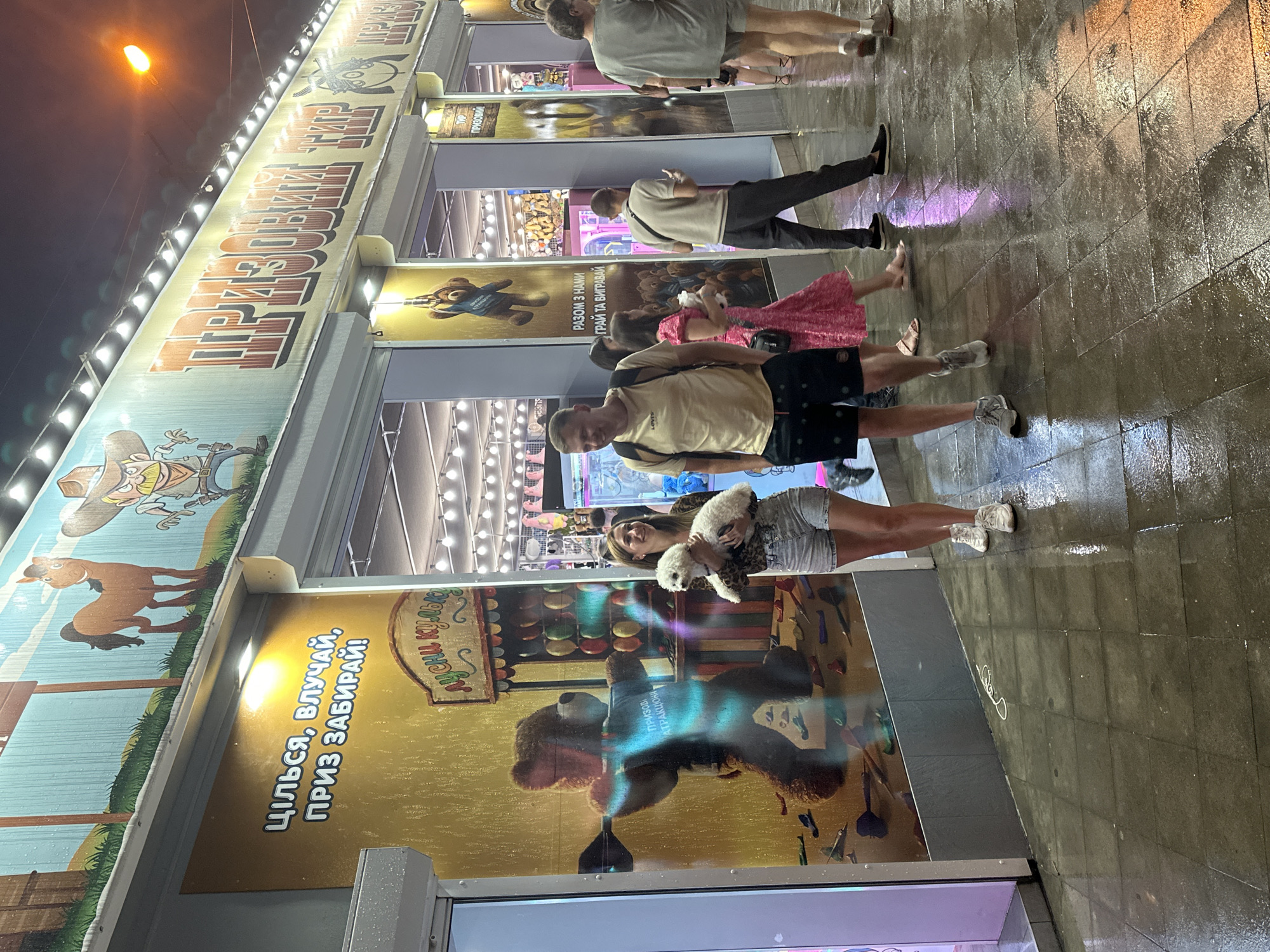 This is a medium shot, taken at night, of a street arcade in Odesa, Ukraine. The arcade is decorated with large murals of cartoon characters and prizes. In the foreground, a woman is holding a small white dog, and a man is walking next to her. They are on a wet pavement, suggesting it has recently rained. Other people are visible in the background, some walking and some standing. The arcade appears to be open, with lights illuminating the interior. The overall atmosphere is lively and festive.