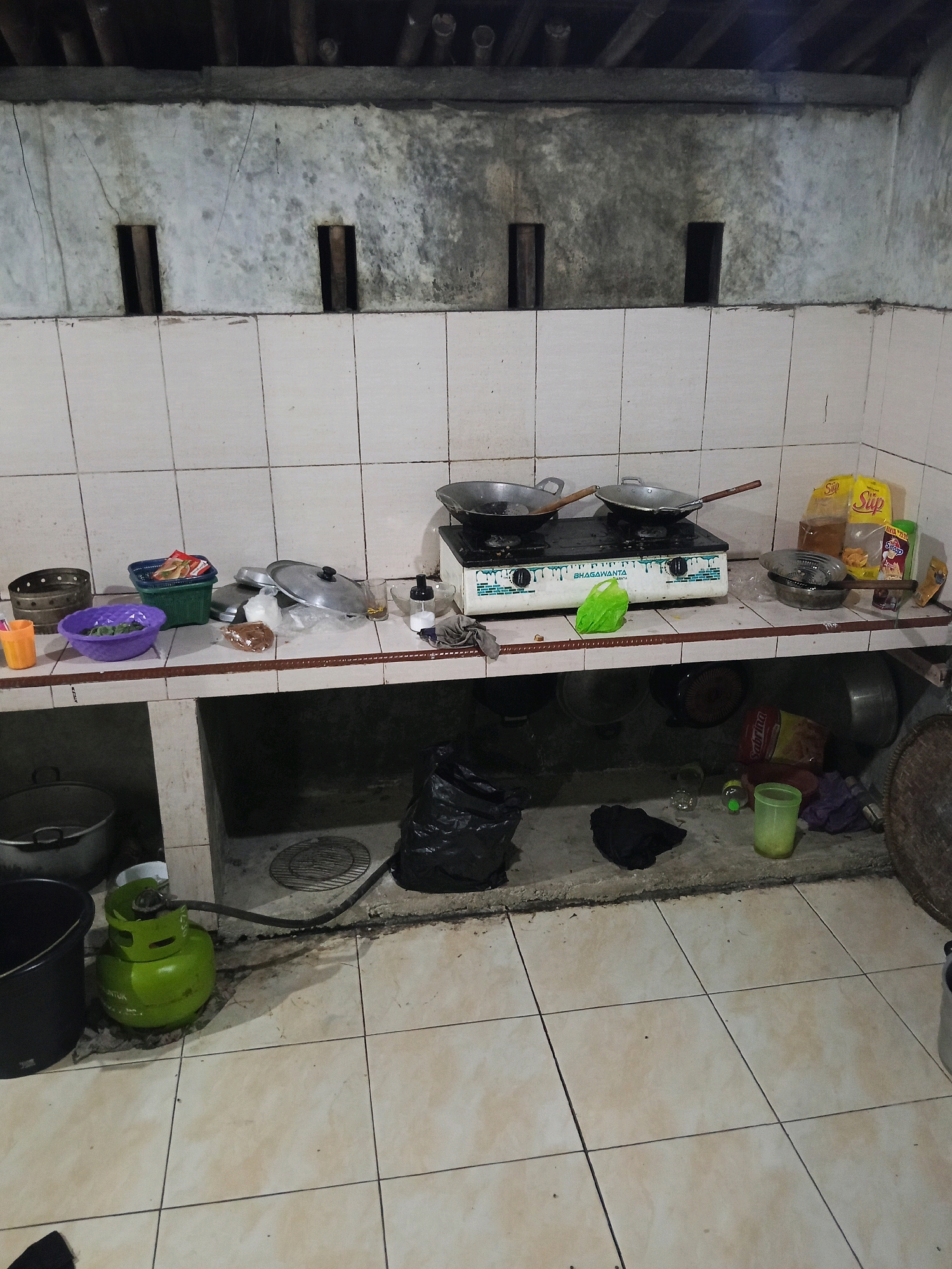 The image depicts an outdoor kitchen area, likely located in Langgongsari, Indonesia. The scene is characterized by a tiled countertop and floor, with a concrete structure beneath the counter. On the countertop, a two-burner gas stove with two pots is visible, along with various containers, a green plastic bag, and packaged food items. Below the counter, there are several large pots and a green gas cylinder connected to the stove via a hose. The wall behind the counter is made of a rough, grey material with two rectangular openings. The overall environment appears somewhat unkempt, with a black plastic bag and other scattered items on the floor.
