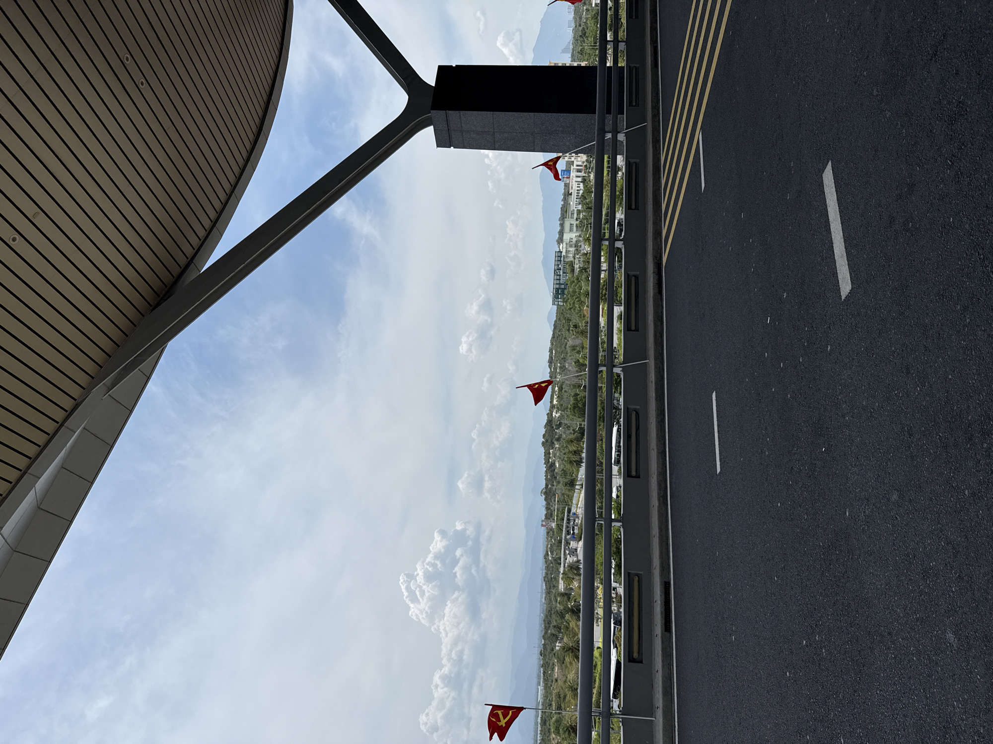 View from Cam Ranh International Airport, Bac Cam Ranh Ward, Vietnam. The image captures a partial view of the airport structure in the foreground, including a curved roof and support beams. Beyond the structure, a landscape is visible, including vegetation, buildings, and distant mountains. Red flags are positioned along a railing. In the foreground, a road with white and yellow painted lines. The sky is cloudy.